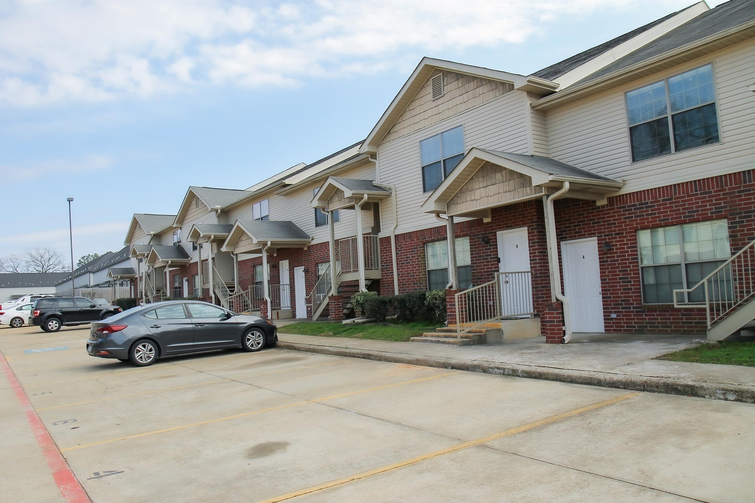 A row of multi-unit townhouses with brick exteriors, featuring stairs leading to white doors. A parked silver car is visible in the foreground, with a paved parking area and a cloudy sky in the background. The scene indicates a suburban residential neighborhood.