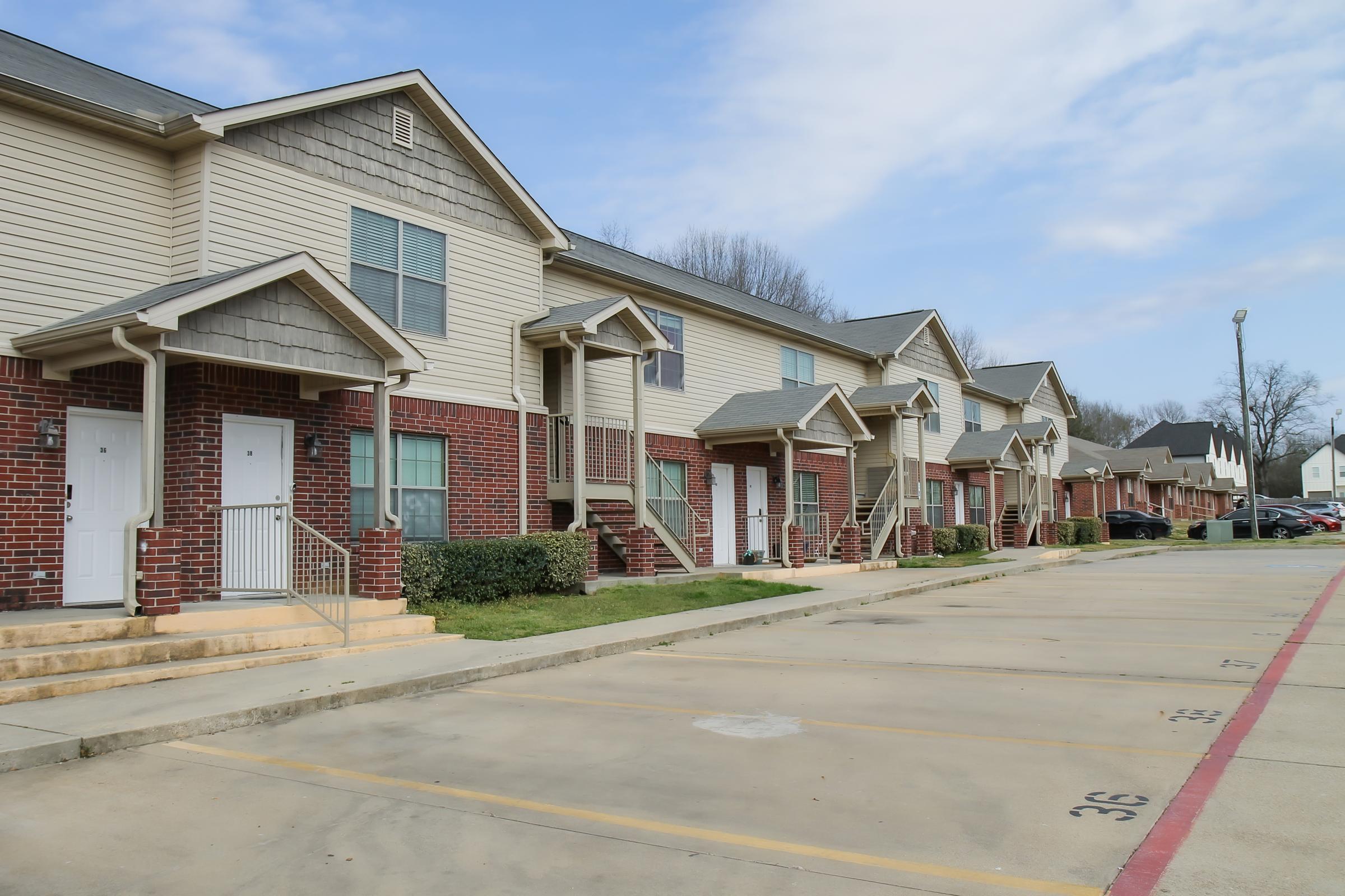 Row of multi-unit residential buildings with a mix of brick and siding exteriors. Each unit features a small porch with stairs leading up to the entrance. The parking lot in front has marked spaces, and greenery is visible along the building's base. The sky is clear with some clouds.