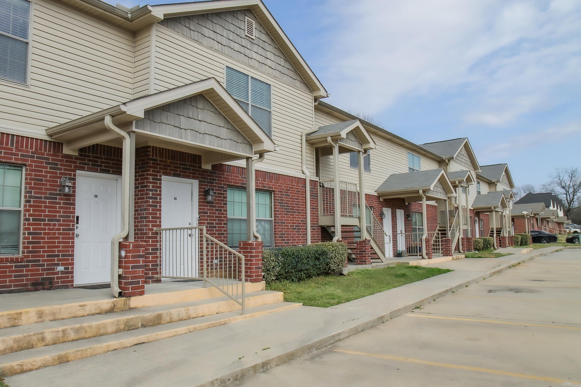 Row of multi-family residential buildings featuring brick and siding exteriors. Each unit has a front door with steps leading up. The area is landscaped with bushes, and a parking lot is visible in front of the buildings under a partly cloudy sky.