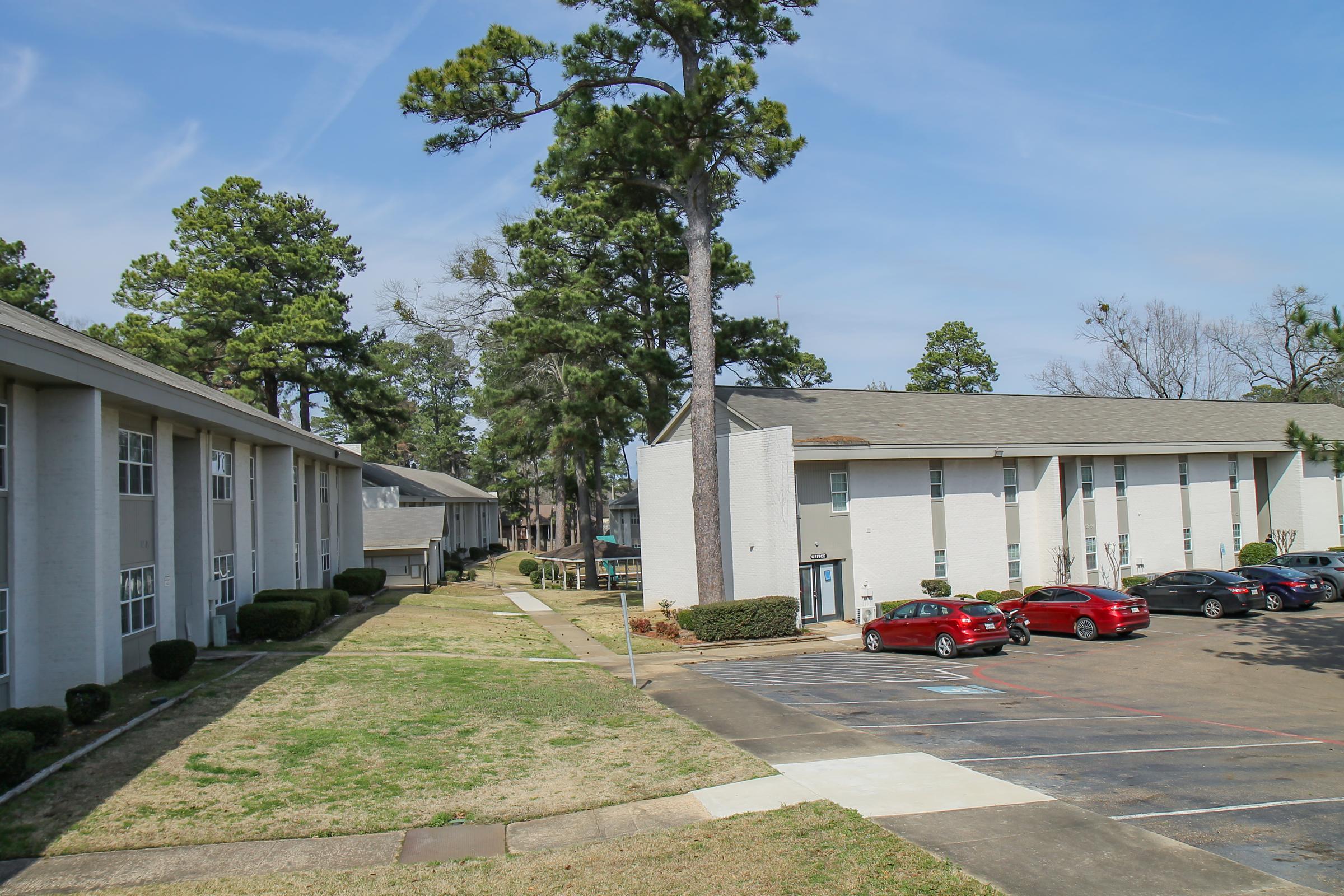 A view of a residential apartment complex surrounded by greenery. Two buildings are visible, one with gray siding and the other white. There are several parked red cars in the parking lot, and well-maintained lawns with trees in the background against a clear blue sky.
