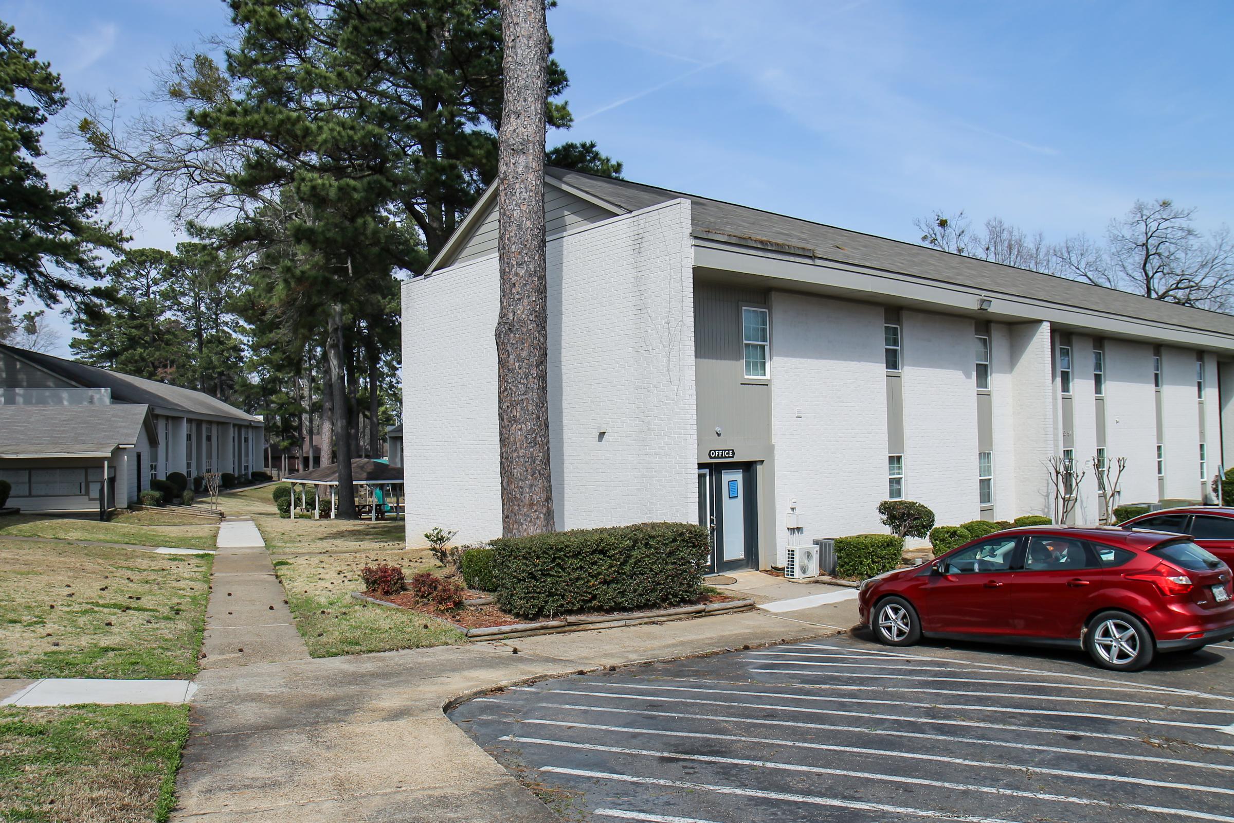A white, multi-story apartment building with a blue door, surrounded by trees and landscaped grass. In the foreground, there is a red car parked in a marked parking area. The scene shows a clear blue sky, indicating a sunny day. The setting appears to be a suburban residential community.