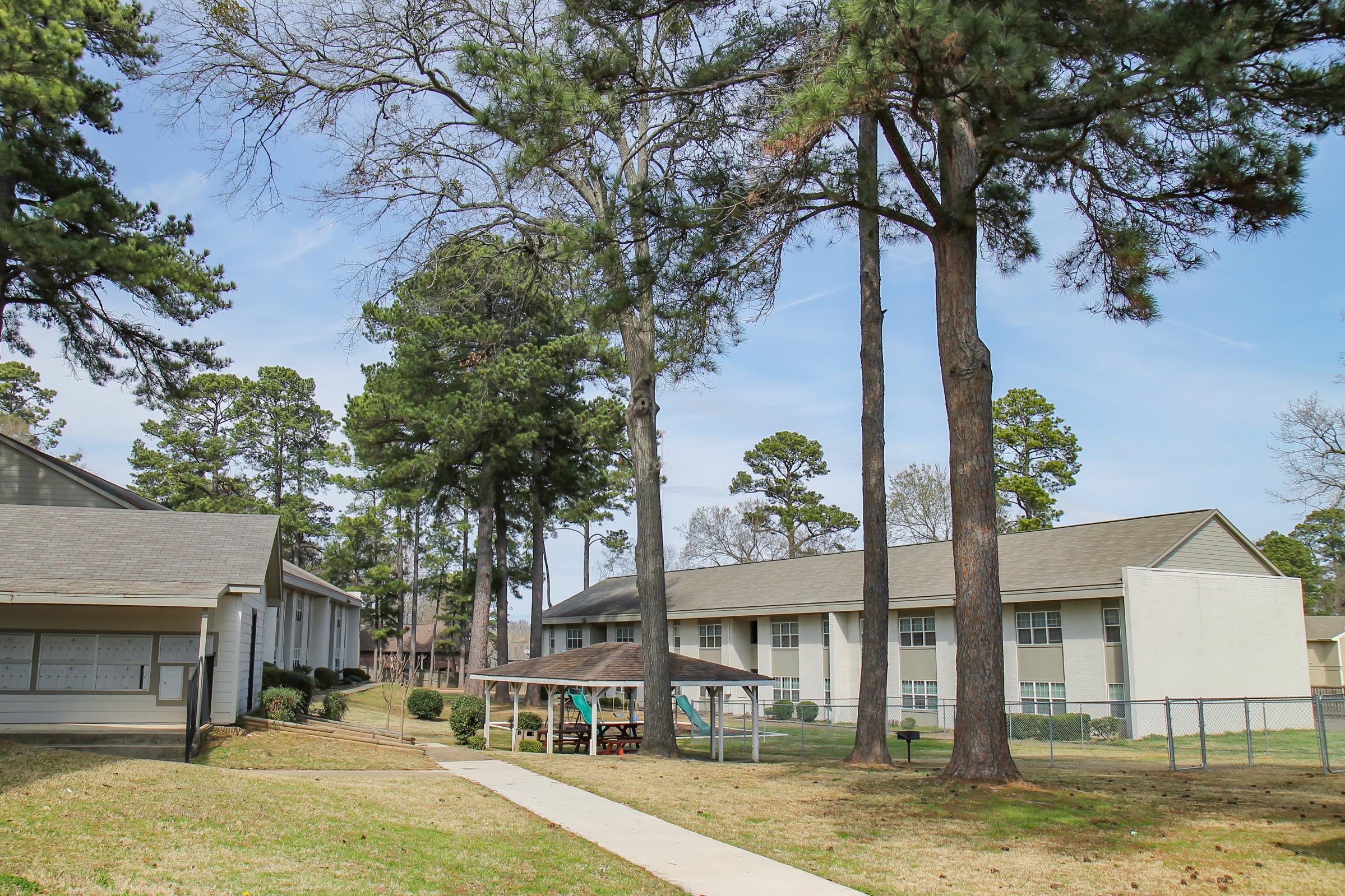 A pathway leads through a grassy area with large trees, flanked by two buildings in a residential setting. One building is two stories high with a lighter facade, while the other is single-story. A wooden structure with a roof is visible in the foreground, suggesting a social or recreational space.