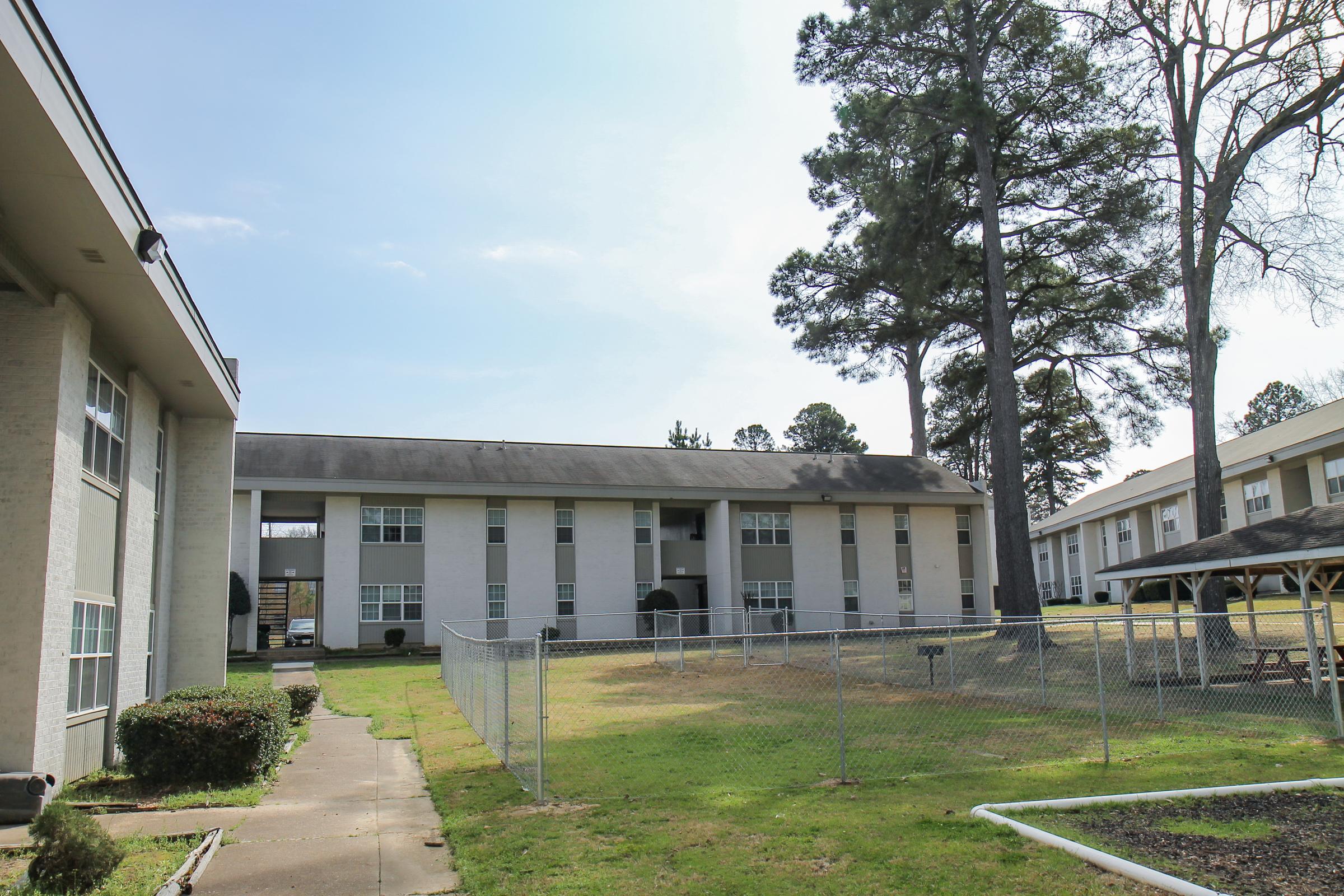 A view of a residential apartment complex featuring two buildings, one with multiple balconies. The scene includes a grassy area with a fence, a picnic gazebo, and tall pine trees. The sky is partly cloudy, creating a serene atmosphere.