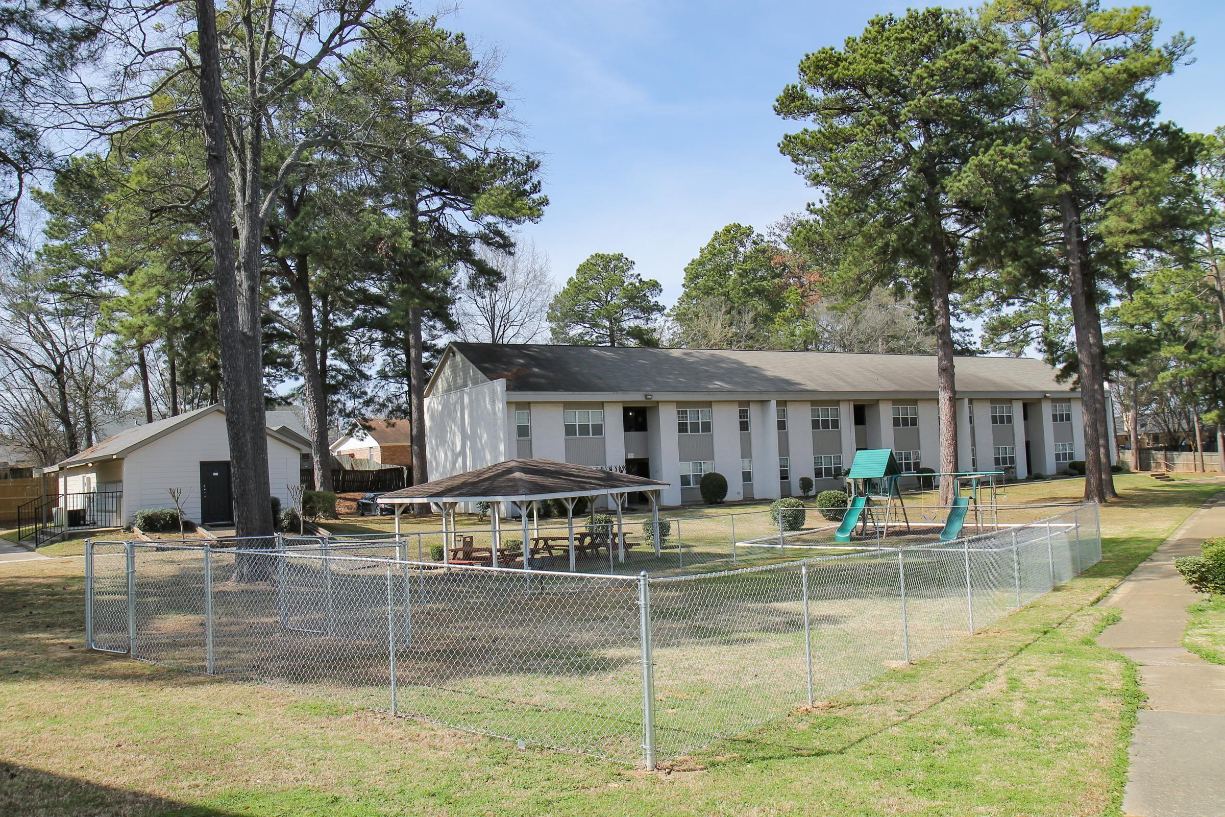View of a multi-unit residential building surrounded by trees, with a fenced area featuring a gazebo, picnic tables, and playground equipment, including a slide and swings. The scene is under a clear blue sky, highlighting a spacious outdoor environment in a residential area.