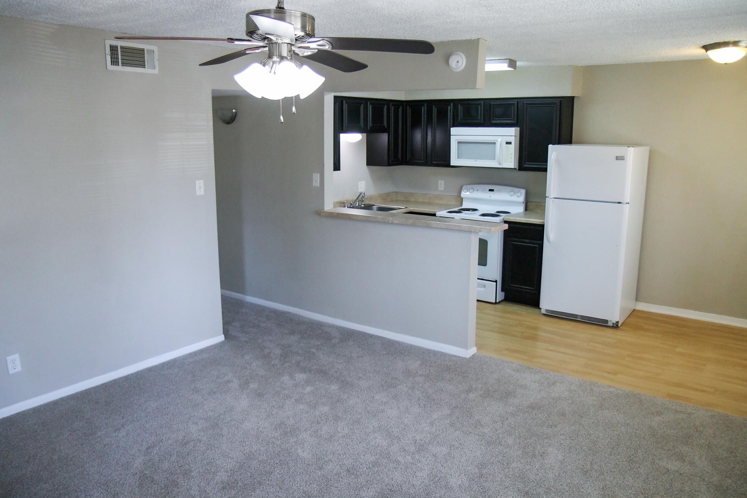 A cozy apartment interior featuring a carpeted living area, a kitchen with dark cabinets, a white stove and refrigerator, and a ceiling fan. The kitchen bar counter separates the living space from the cooking area, creating an open layout. Natural light from a nearby window brightens the room.