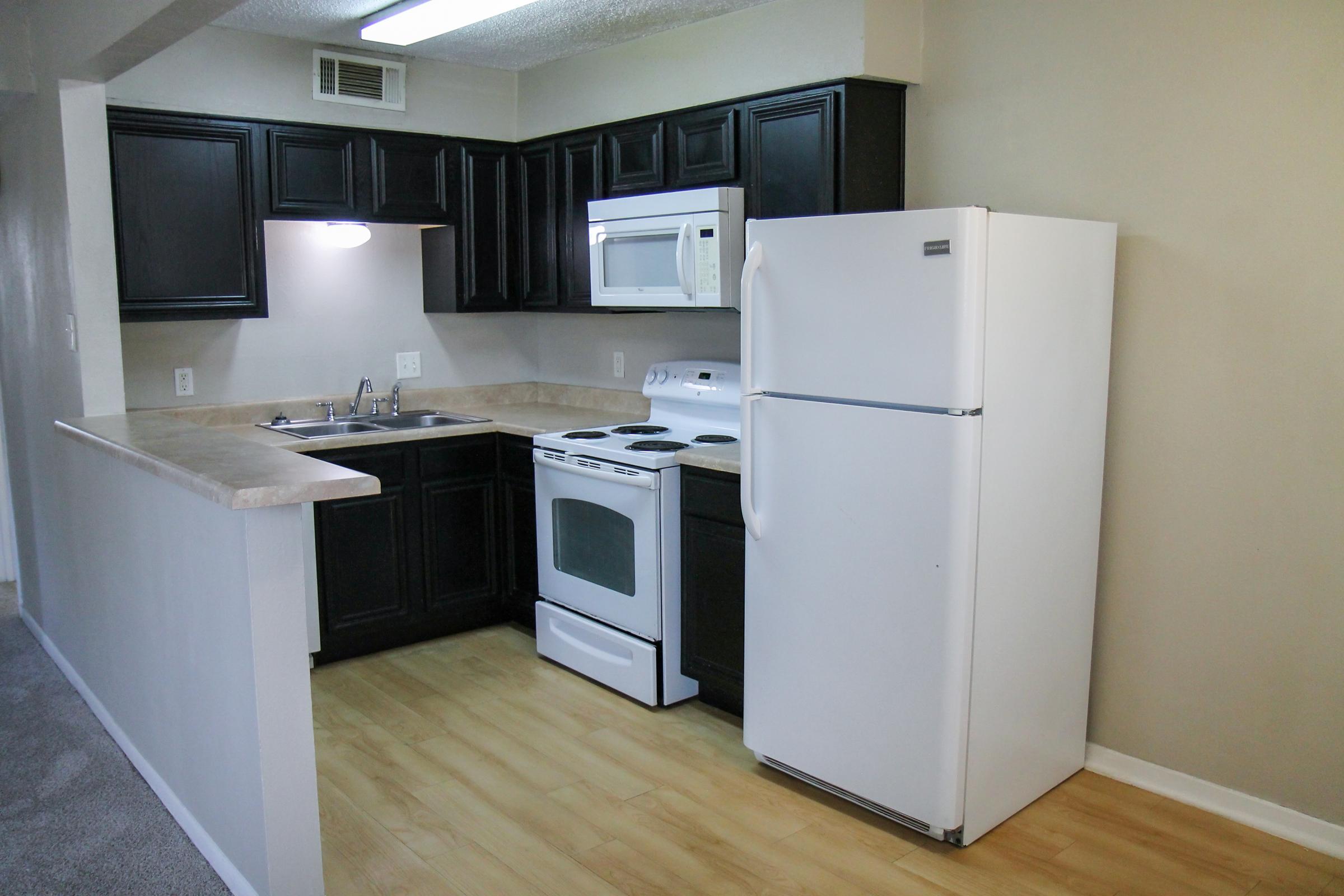 Modern kitchen featuring black cabinetry, a light counter, a white stove and refrigerator, and a microwave. The space has a light-colored wall and wooden flooring, creating a clean and inviting atmosphere.