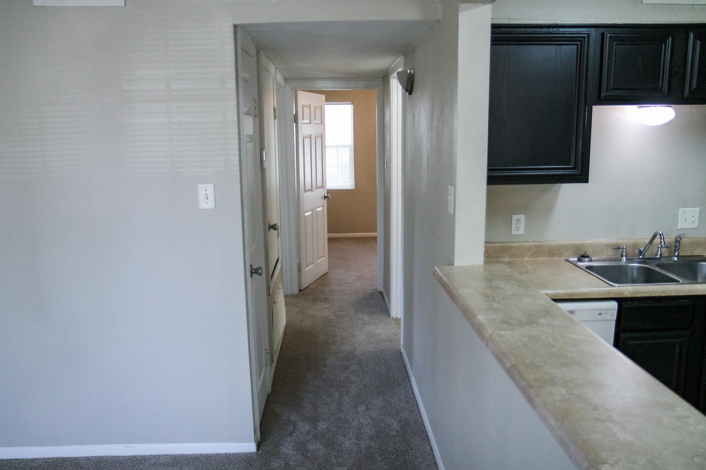 A view from a kitchen leading to a hallway with light-colored walls and carpet. A doorway at the end of the hallway opens to a well-lit room, while the kitchen features dark cabinets, a countertop, and a sink.