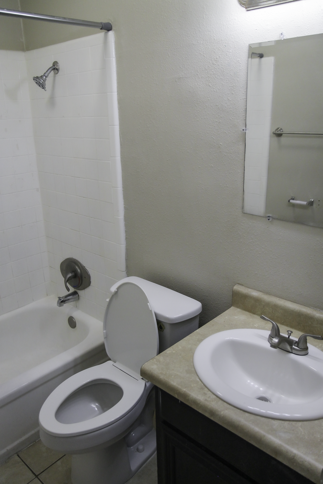 A small bathroom featuring a white bathtub with a showerhead, a toilet, and a sink with a faucet. The sink area has a light-colored countertop, and there's a mirror above the sink. The walls are painted a light color, and the floor is tiled.