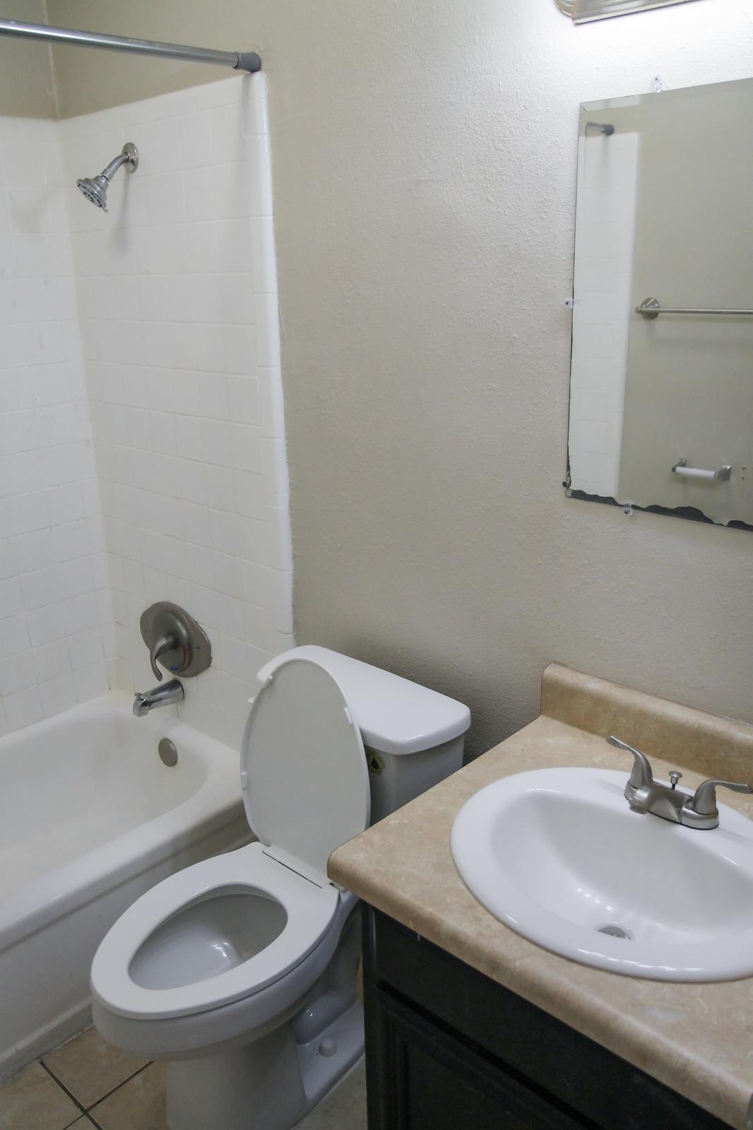 A simple bathroom featuring a bathtub with a shower, a toilet, and a sink with a mirror above it. The walls are painted a light color, and the floor is tiled. The overall design is functional and modest.