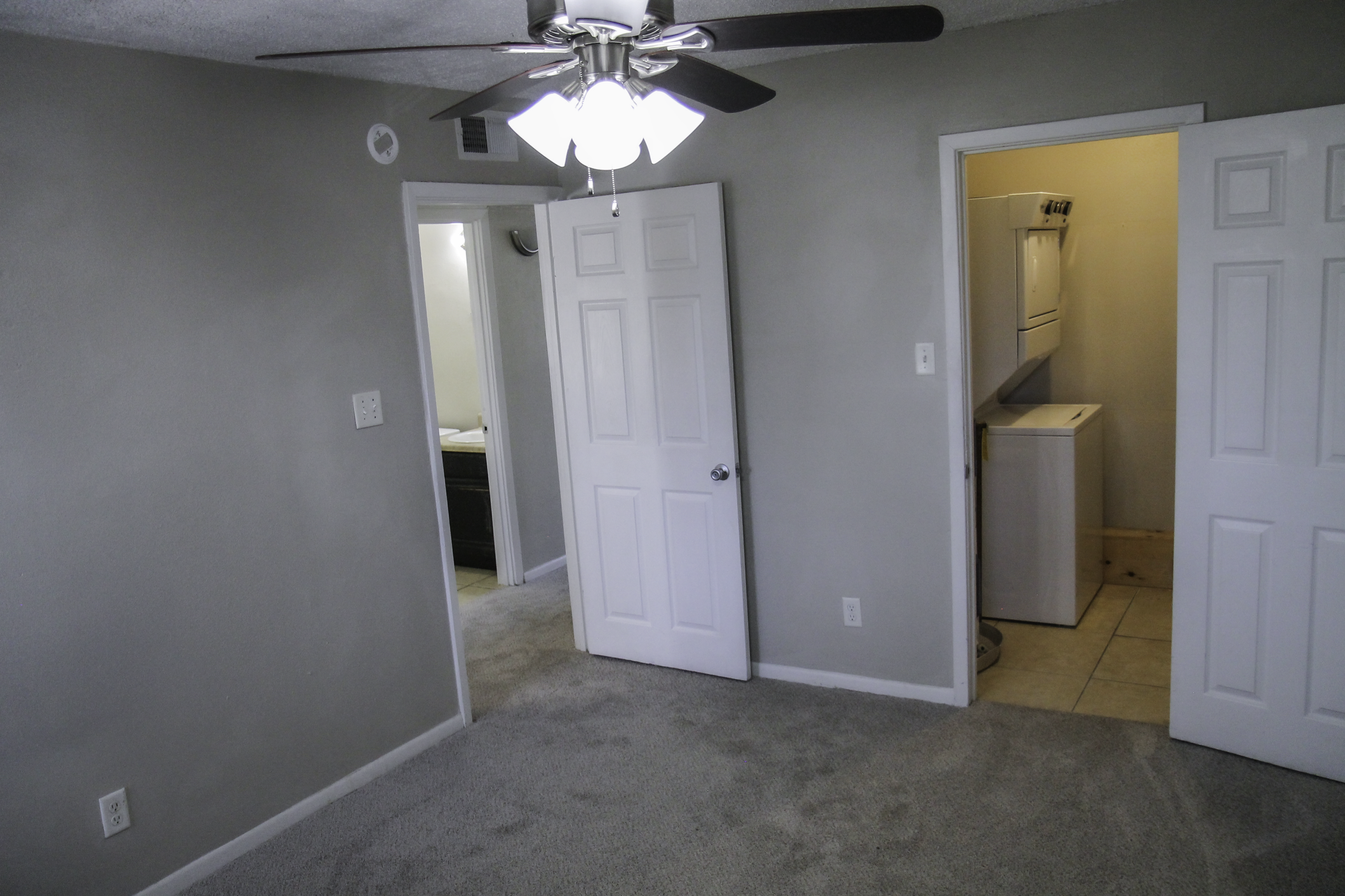 A well-lit room with light gray walls and carpet. A ceiling fan is visible, and there are two doors: one leading to a laundry area and the other to an adjacent room. The overall atmosphere is clean and minimalistic, suitable for a living space.