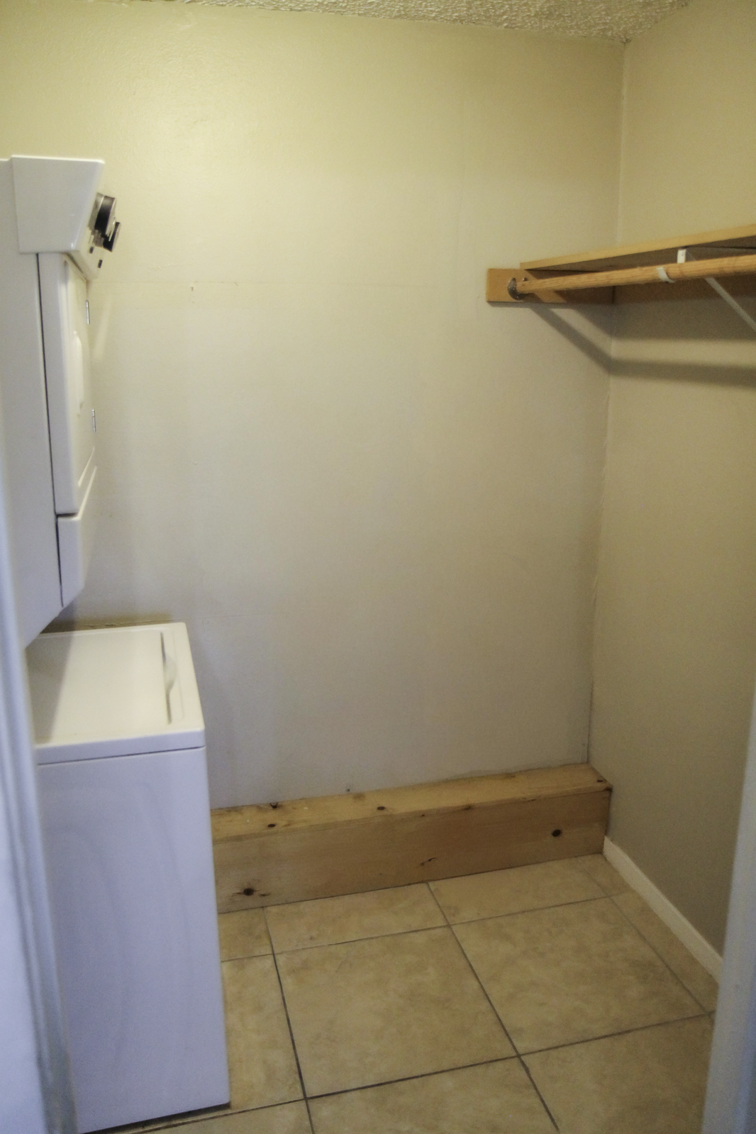 A small laundry room featuring a stacked washer and dryer on the left, and a wooden shelf along the wall on the right. The walls are painted a light color, and the floor is tiled. The space appears organized and functional, suitable for laundry tasks.