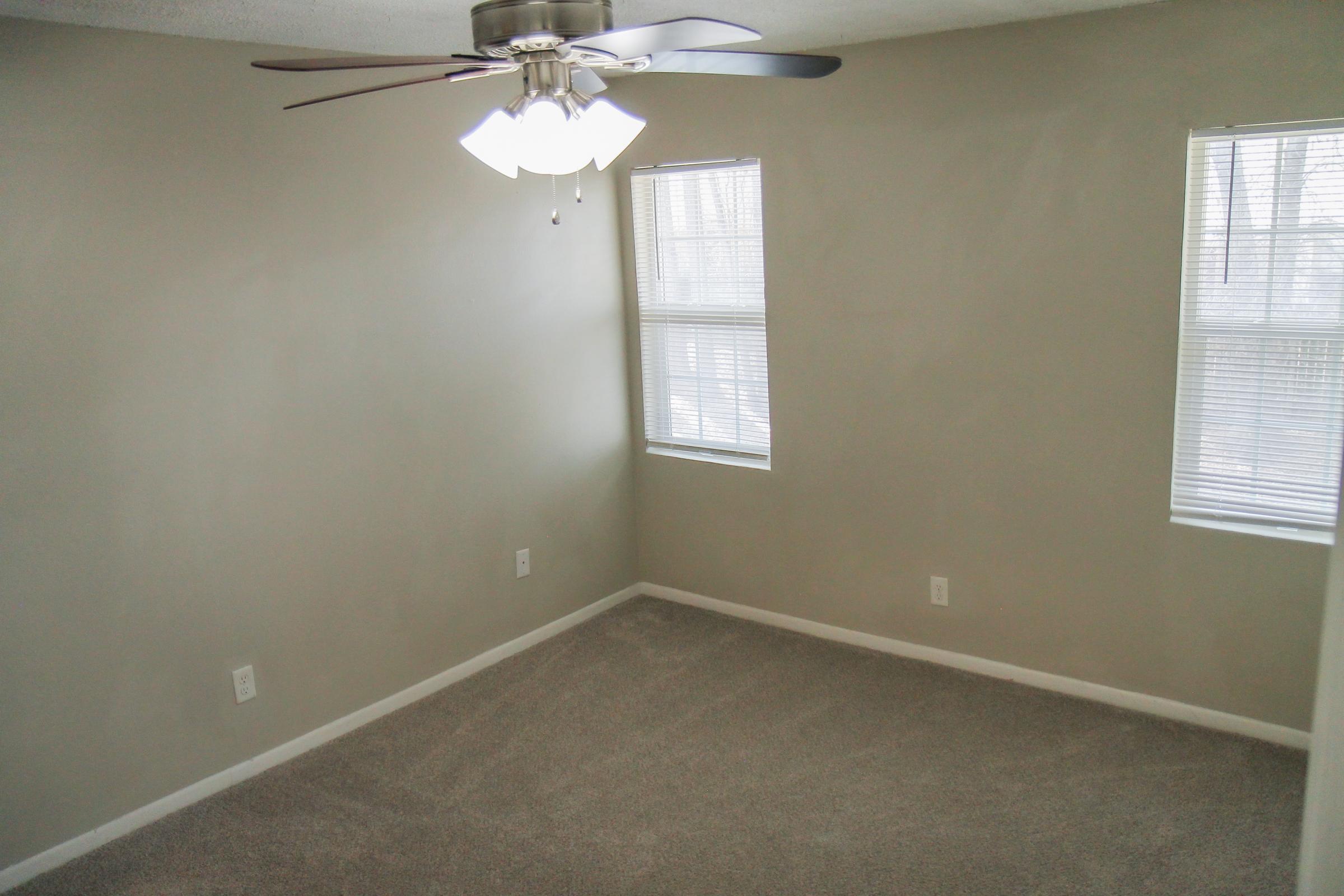 A well-lit, empty room featuring light gray walls and carpet. It has two windows with blinds letting in natural light, and a ceiling fan with four lights. The space appears clean and spacious, ideal for various purposes such as a bedroom or office.