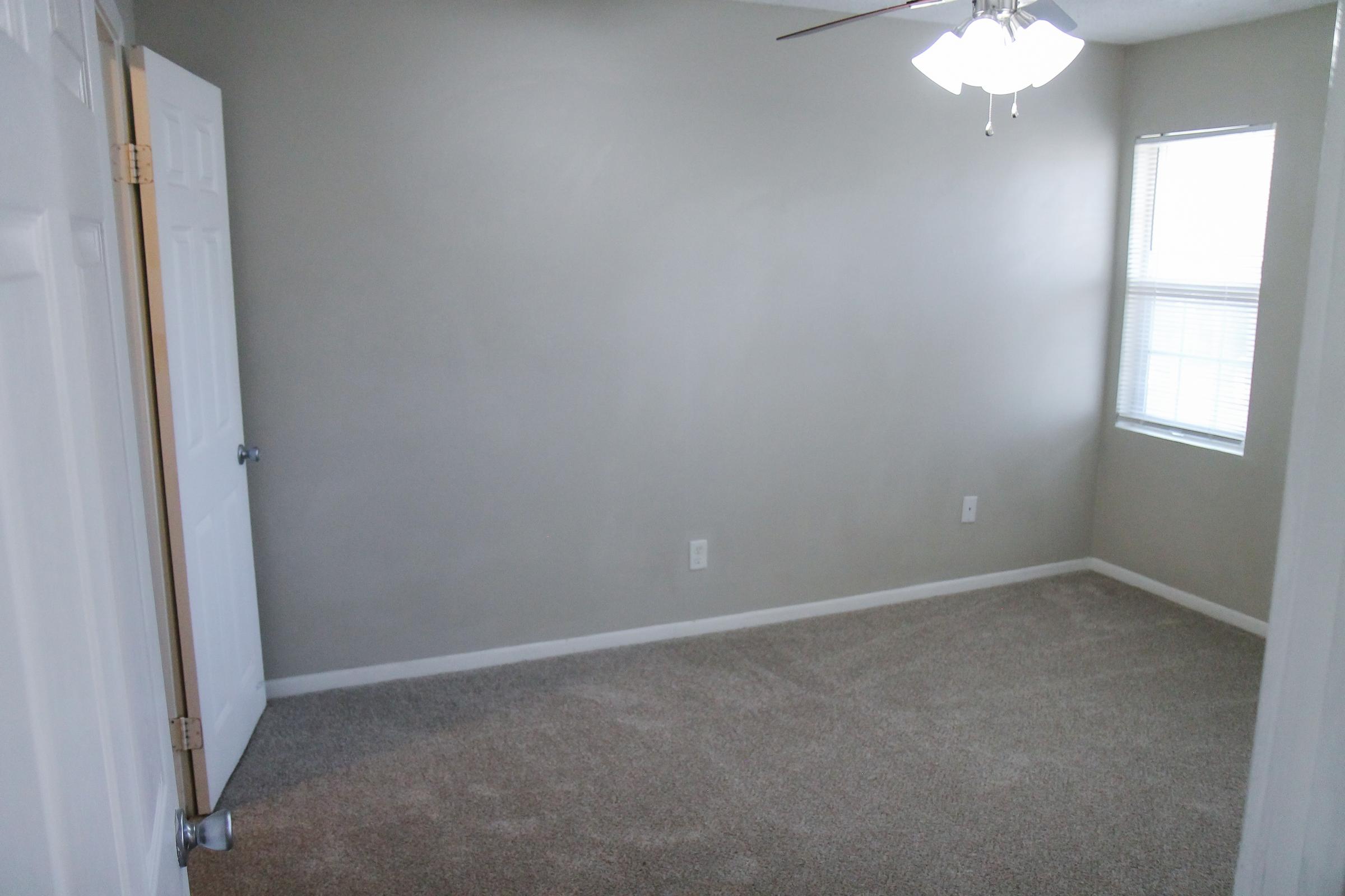 A small, empty room with light gray walls and beige carpet. There is a ceiling fan with three blades, and a single window with blinds allowing natural light to enter. The door is partially open, leading to another space outside the room. The overall atmosphere is clean and uncluttered.