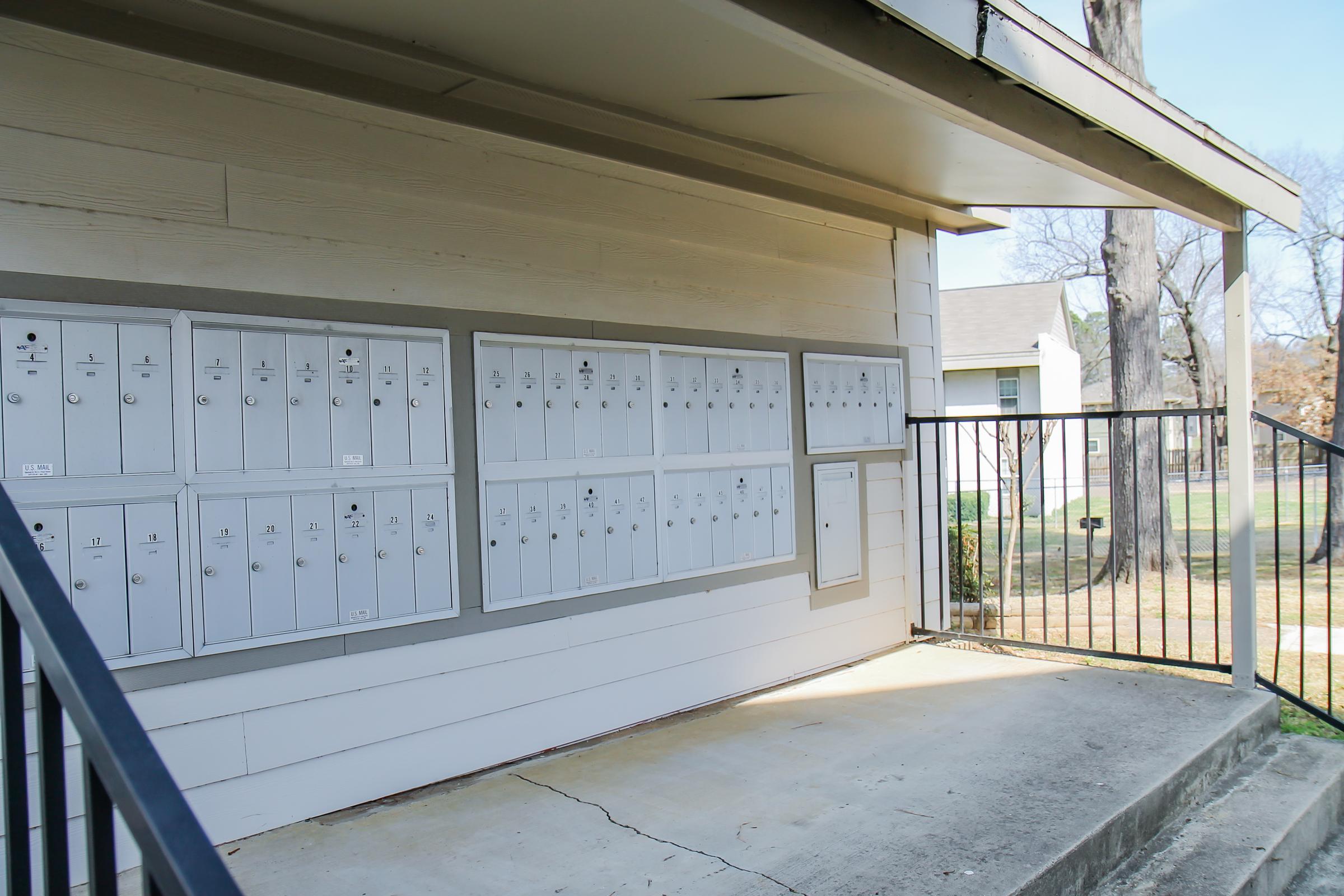A row of white mailboxes attached to the side of a building, situated under a covered area. The mailboxes are organized in a grid pattern, with a few empty spaces. A metal railing is visible in the foreground, and some greenery can be seen in the background, indicating an outdoor setting.