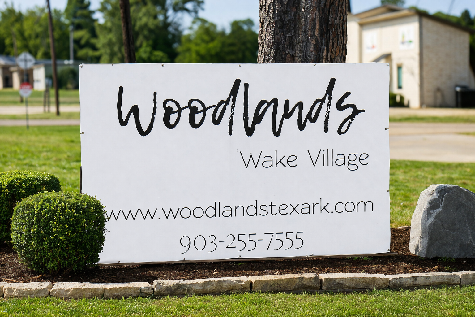 Sign for Woodlands in Wake Village, displaying the website and contact number. The sign is white with black text, set against a green grassy landscape with trees and shrubs nearby.