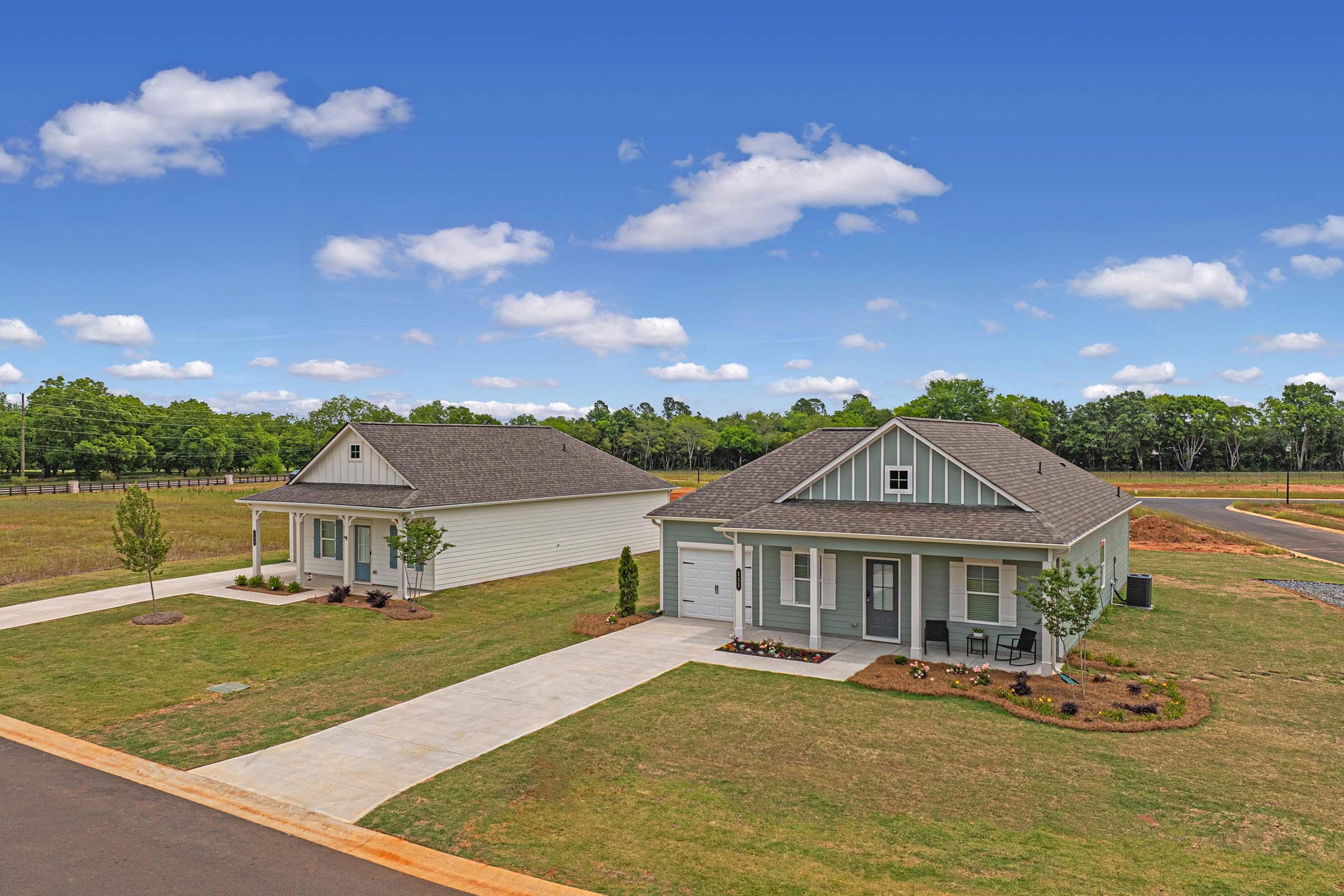 Two single-story houses with manicured lawns and driveways, set against a clear blue sky with scattered clouds. The houses feature light-colored exteriors and dark roof shingles. Surrounding the homes is open land with green grass and trees in the background.