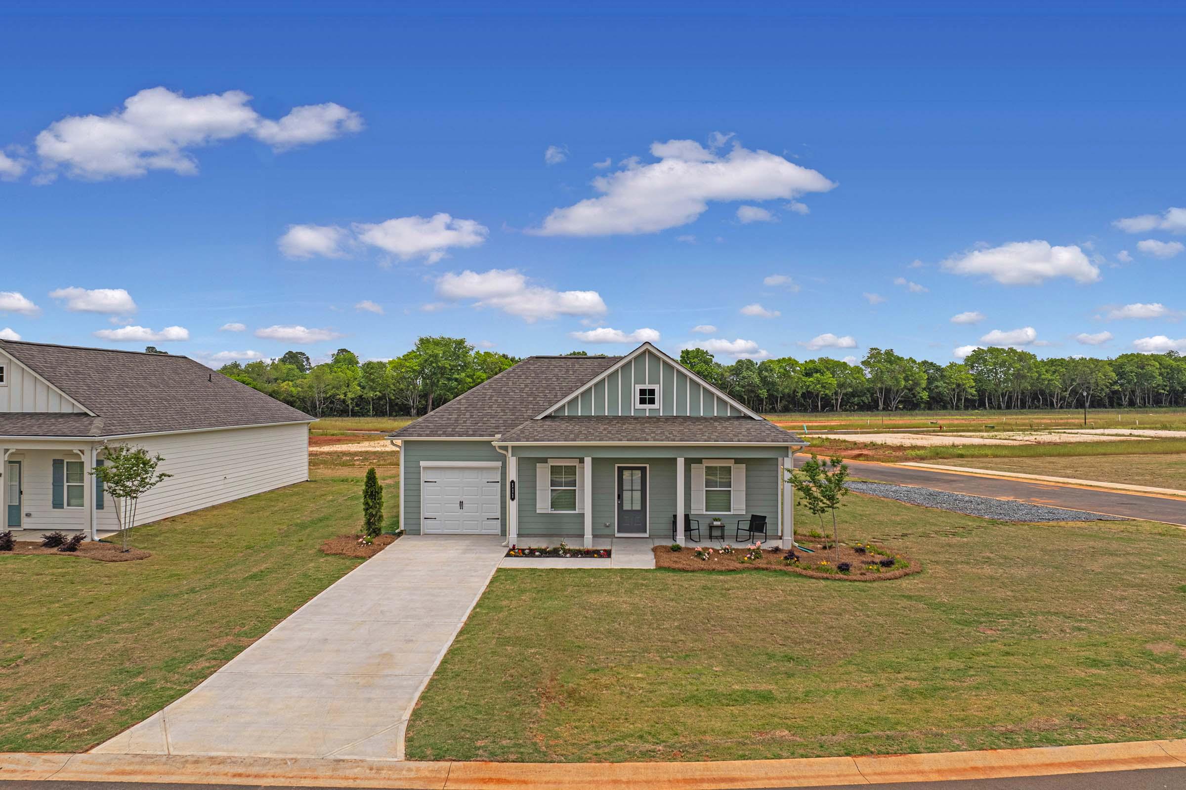 A single-story light blue house with a front porch, situated on a grassy lot. It features a driveway and is surrounded by small shrubs. The background includes trees and an open field under a bright blue sky with a few clouds.