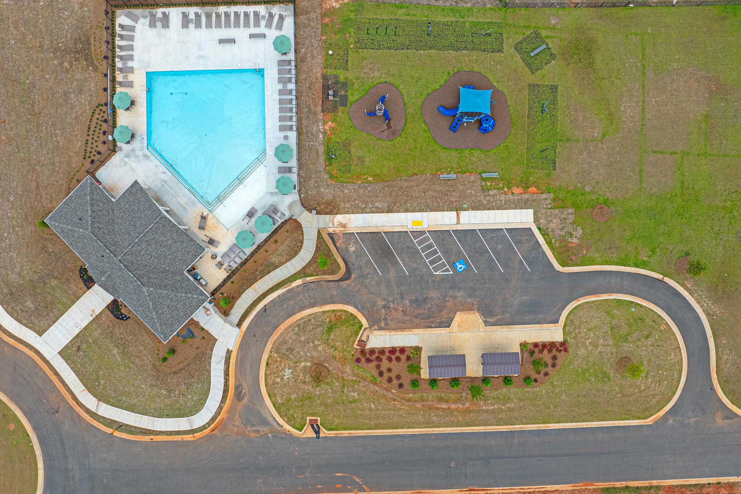 An aerial view of a residential area featuring a swimming pool, playground, parking area, and neatly landscaped surroundings. The pool is bright blue and flanked by lounge chairs, while the playground includes structures for children. A curved road and pathways add to the community layout.