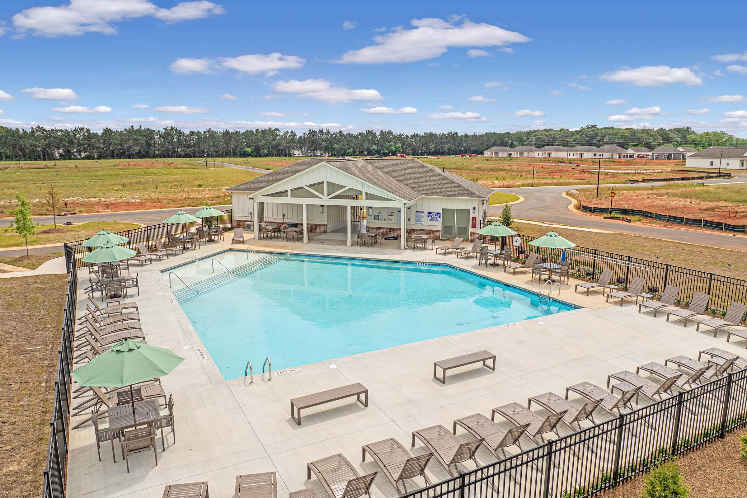 Aerial view of a community pool area featuring a rectangular blue swimming pool surrounded by lounge chairs and green umbrellas. There is a clubhouse nearby, and the landscape includes grassy areas and distant residential buildings under a partly cloudy sky.