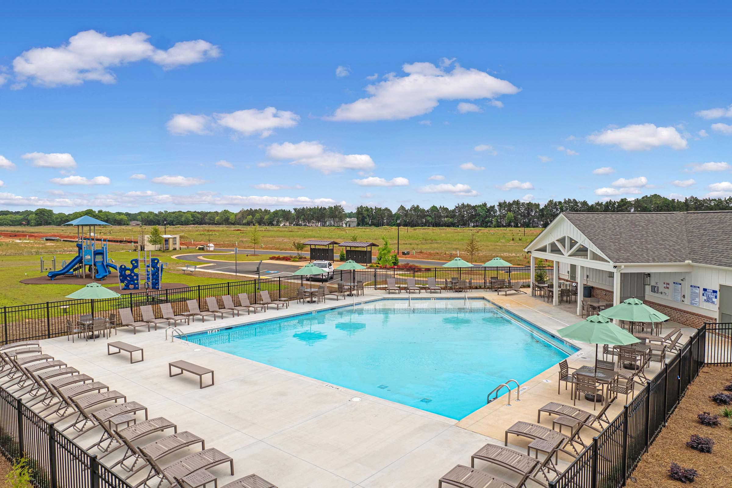 A clear blue swimming pool surrounded by lounge chairs and green umbrellas, with a grassy field and playground equipment in the background. A modern building with a covered area is nearby, under a bright blue sky with fluffy white clouds.