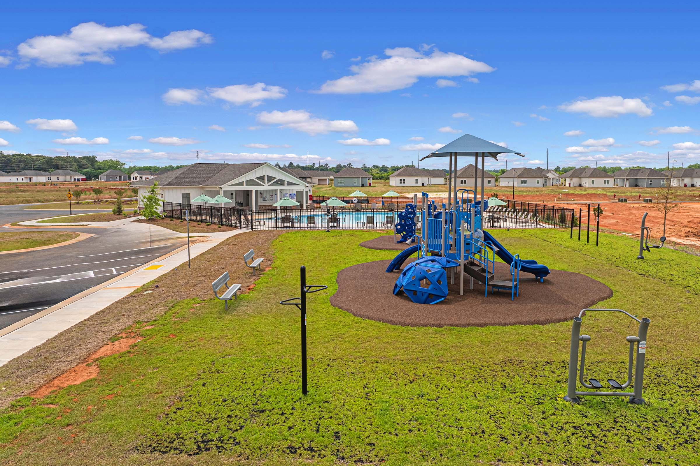 A brightly colored playground featuring a blue play structure with slides and climbing elements, surrounded by green grass. Nearby, there are benches and outdoor fitness equipment. In the background, a community pool and residential buildings are visible under a clear blue sky with clouds.