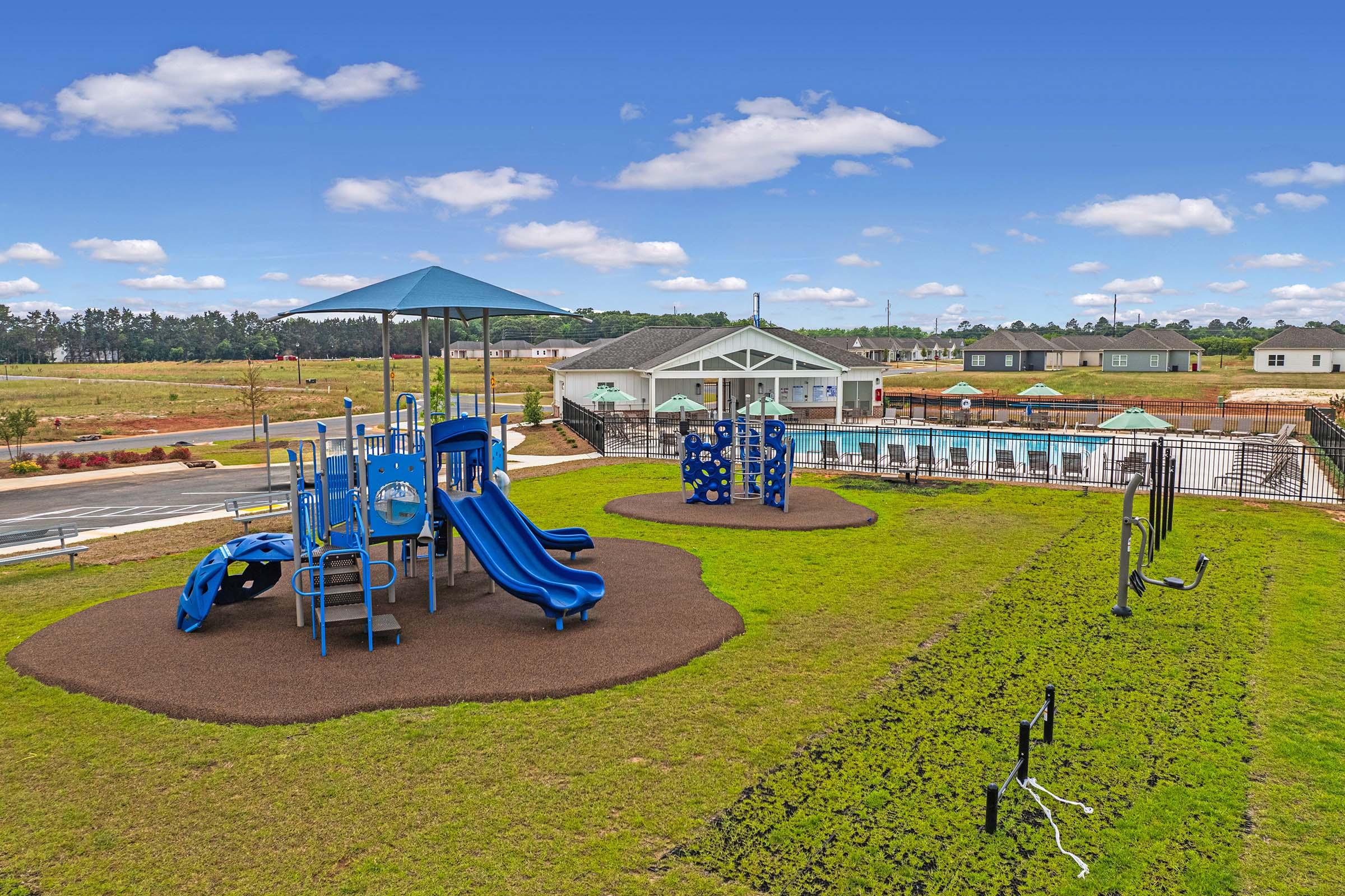 Playground featuring blue slides and climbing structures on a grassy area. In the background, there is a swimming pool and several residential buildings. The sky is clear with scattered clouds, providing a bright and sunny atmosphere.
