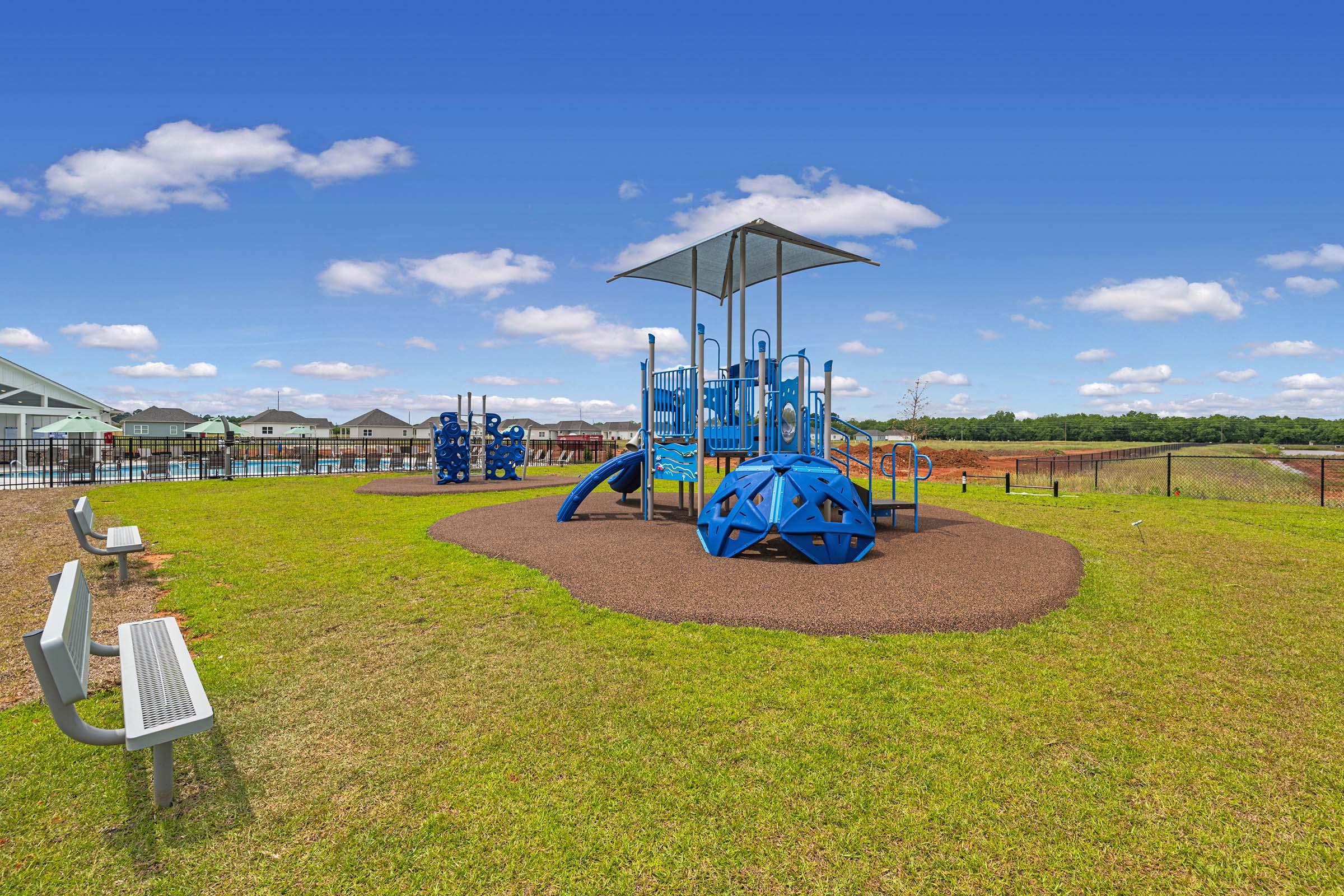 A vibrant playground featuring blue structures, including slides and climbing apparatus, set on a grassy area. Nearby, there are benches for seating, and a clear blue sky with scattered clouds overhead. In the background, modern buildings and a fence can be seen, indicating the playground is part of a community setup.