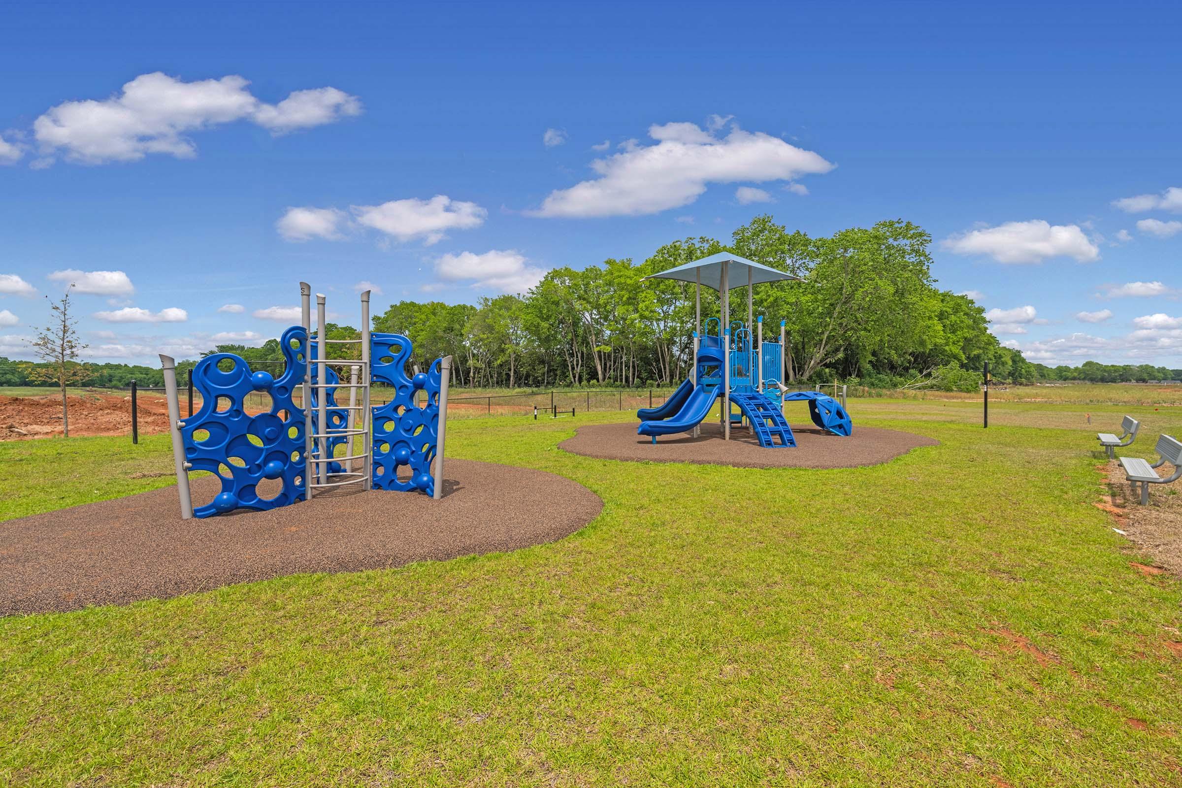 A colorful blue playground structure featuring climbing elements and a slide, surrounded by a grassy area. In the background, there are trees and a clear blue sky with a few clouds. Benches are visible nearby, indicating a space for relaxation.
