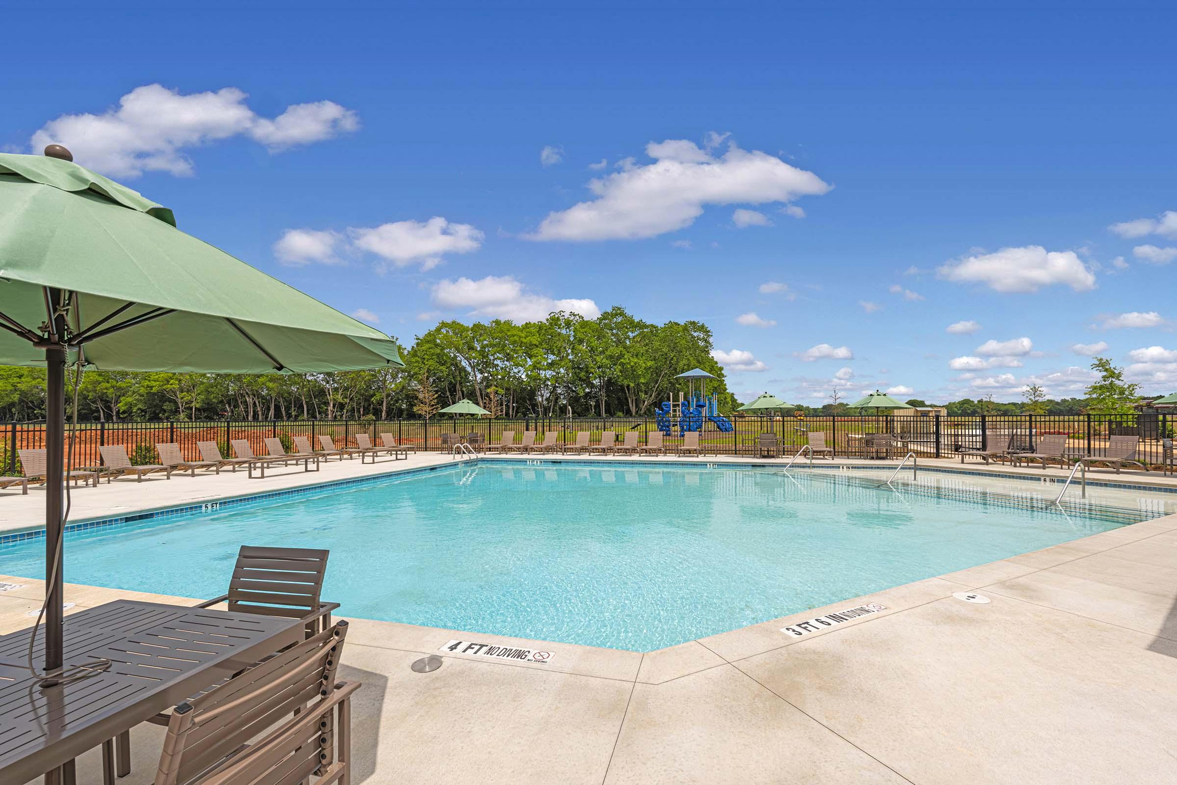 A clear blue swimming pool surrounded by lounge chairs and green umbrellas, with a fenced area and grassy landscape in the background. The sky is bright with a few fluffy clouds, and there is a playground visible in the distance.