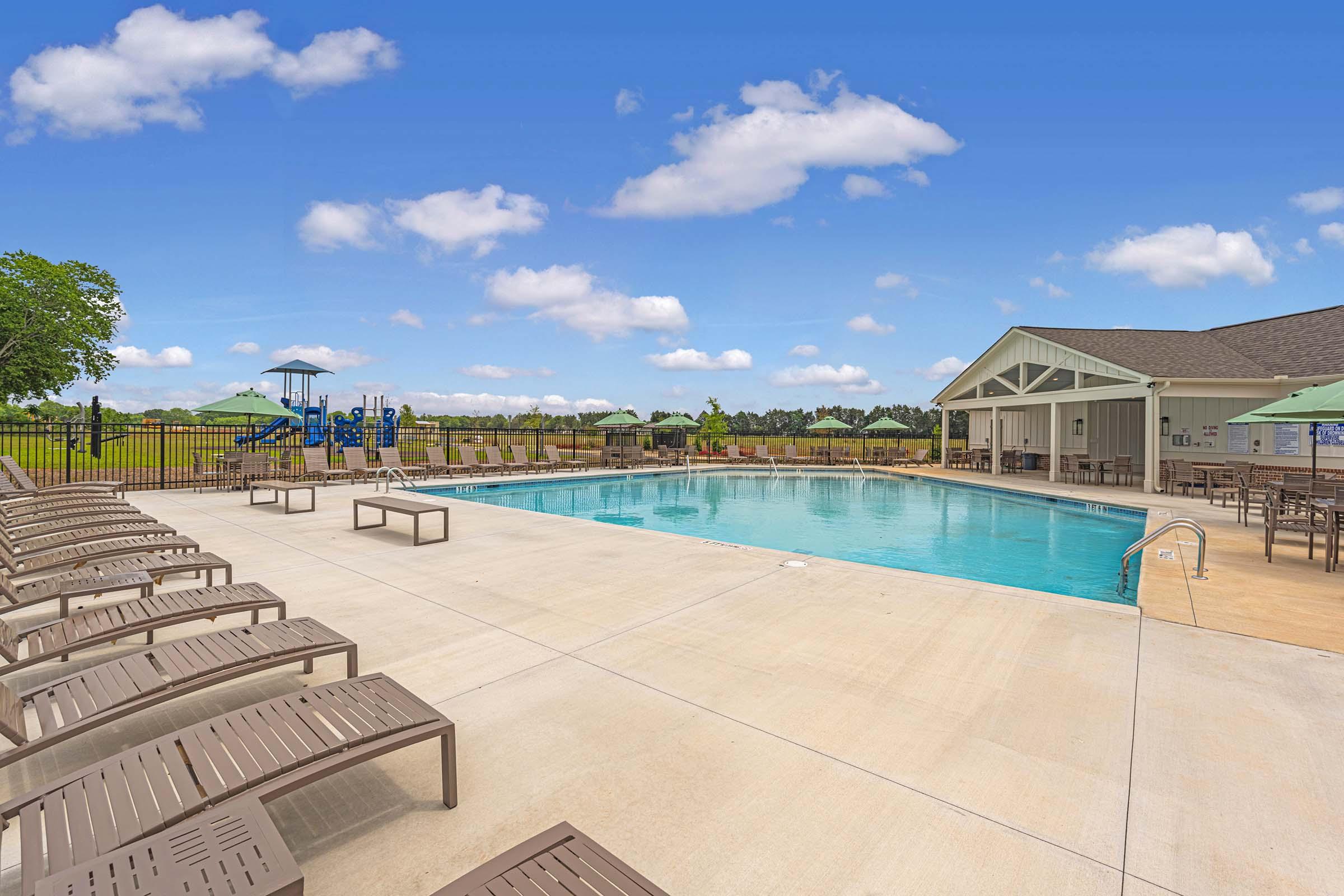 A bright, inviting outdoor pool area featuring lounge chairs, a large swimming pool, and a nearby playground. The landscape includes green grass and a clear blue sky with fluffy white clouds. A clubhouse is visible in the background, providing a welcoming atmosphere for relaxation and recreation.