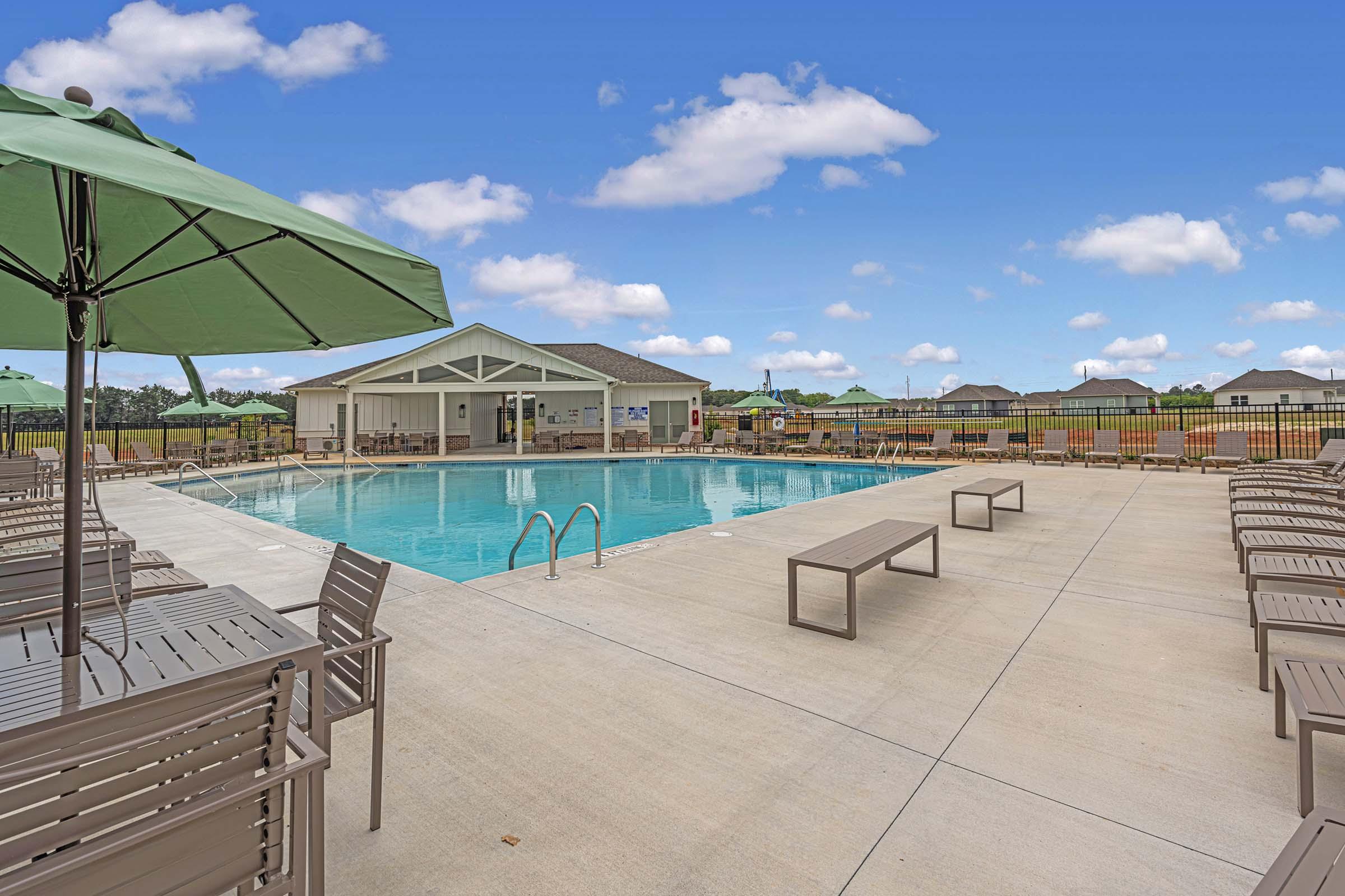 A serene outdoor swimming pool surrounded by lounge chairs and green umbrellas. In the background, there is a clubhouse and landscaped views under a clear blue sky with fluffy white clouds. The area is designed for relaxation and recreation.