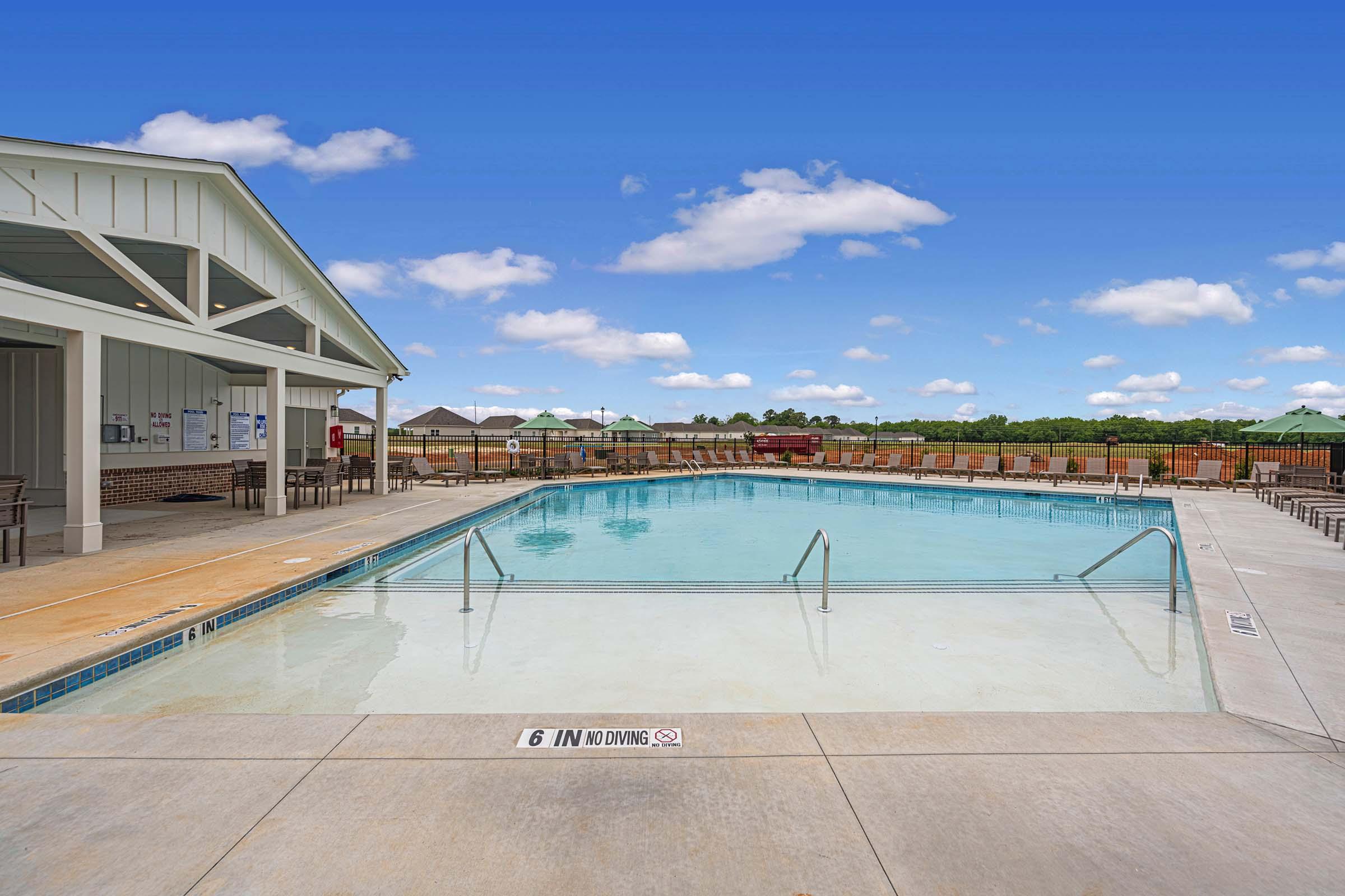 A clear swimming pool with no diving sign, surrounded by a concrete deck. Loungers and an outdoor pavilion are visible, with a backdrop of green trees under a bright blue sky with fluffy clouds. The scene conveys a relaxing outdoor atmosphere.