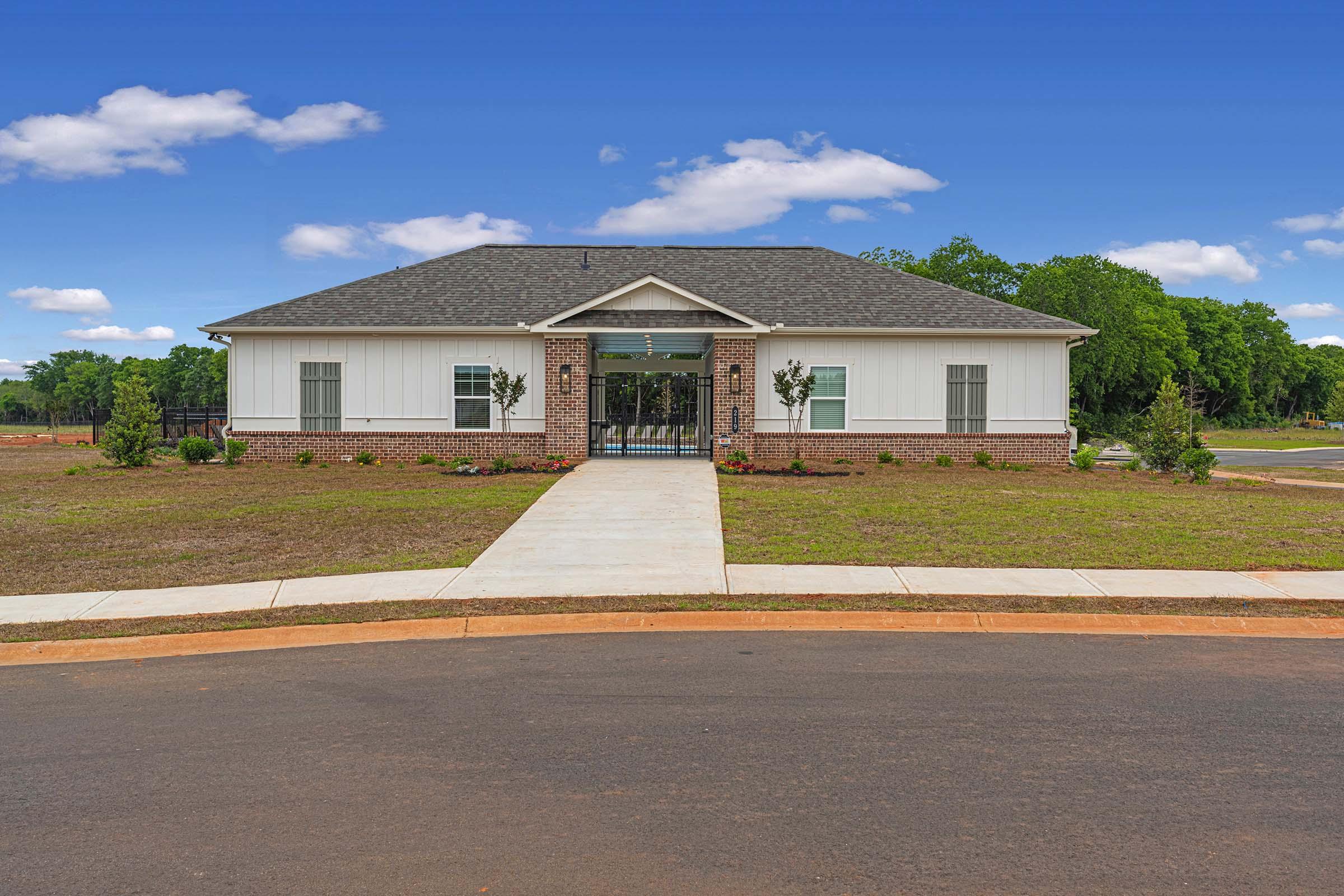 A modern single-story building with a white and brick facade, set against a blue sky with fluffy clouds. A paved walkway leads to the entrance, flanked by small shrubs and landscaping. The area is mostly grassy, with a curved road in the foreground, suggesting a residential or community setting.