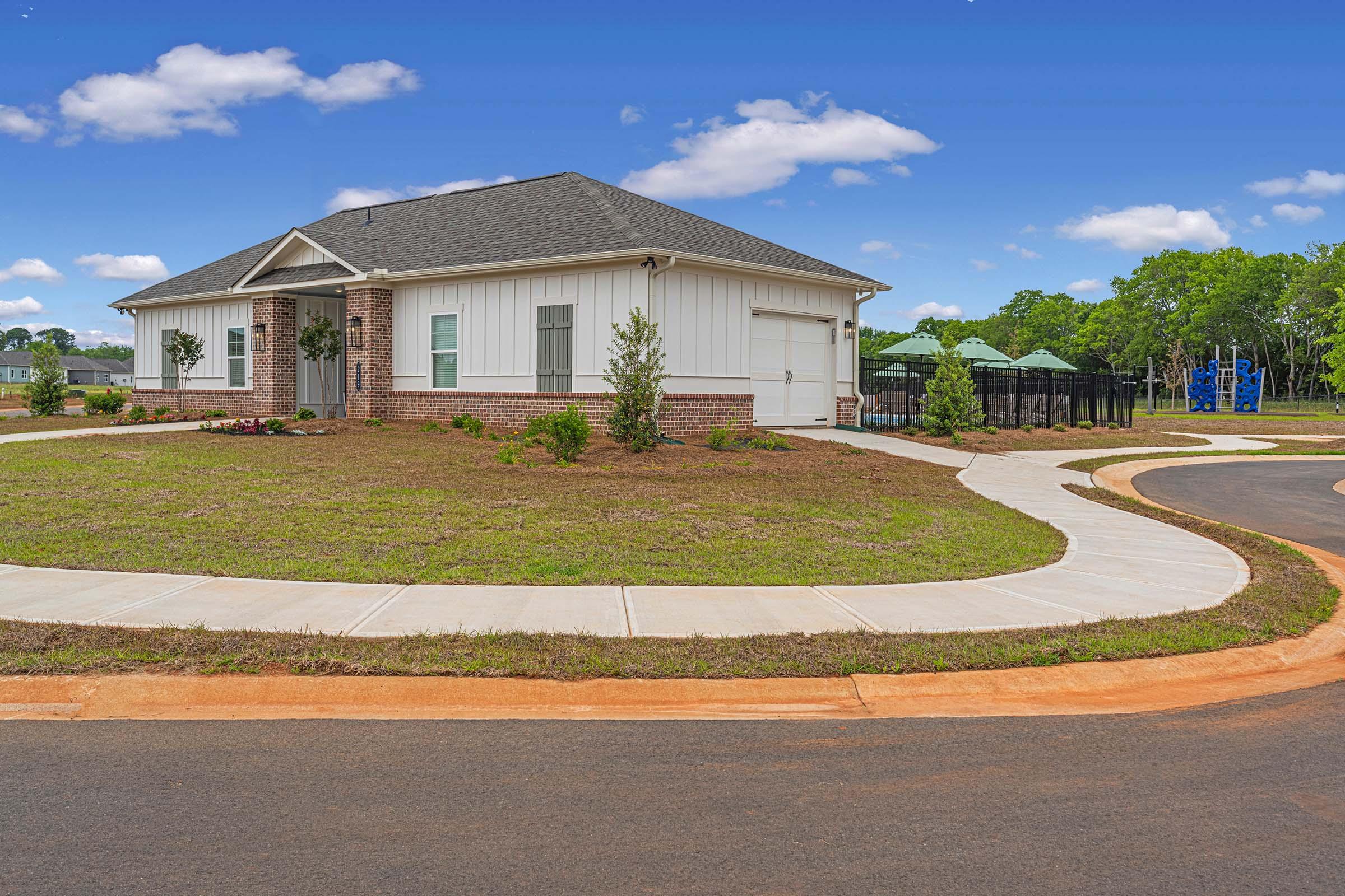 A modern single-story house with a light-colored exterior and a dark roof, situated on a corner lot. The lawn is freshly landscaped with small shrubs and flowers. A curved sidewalk leads to the front of the house, and playground equipment can be seen in the background, indicating a family-friendly neighborhood.