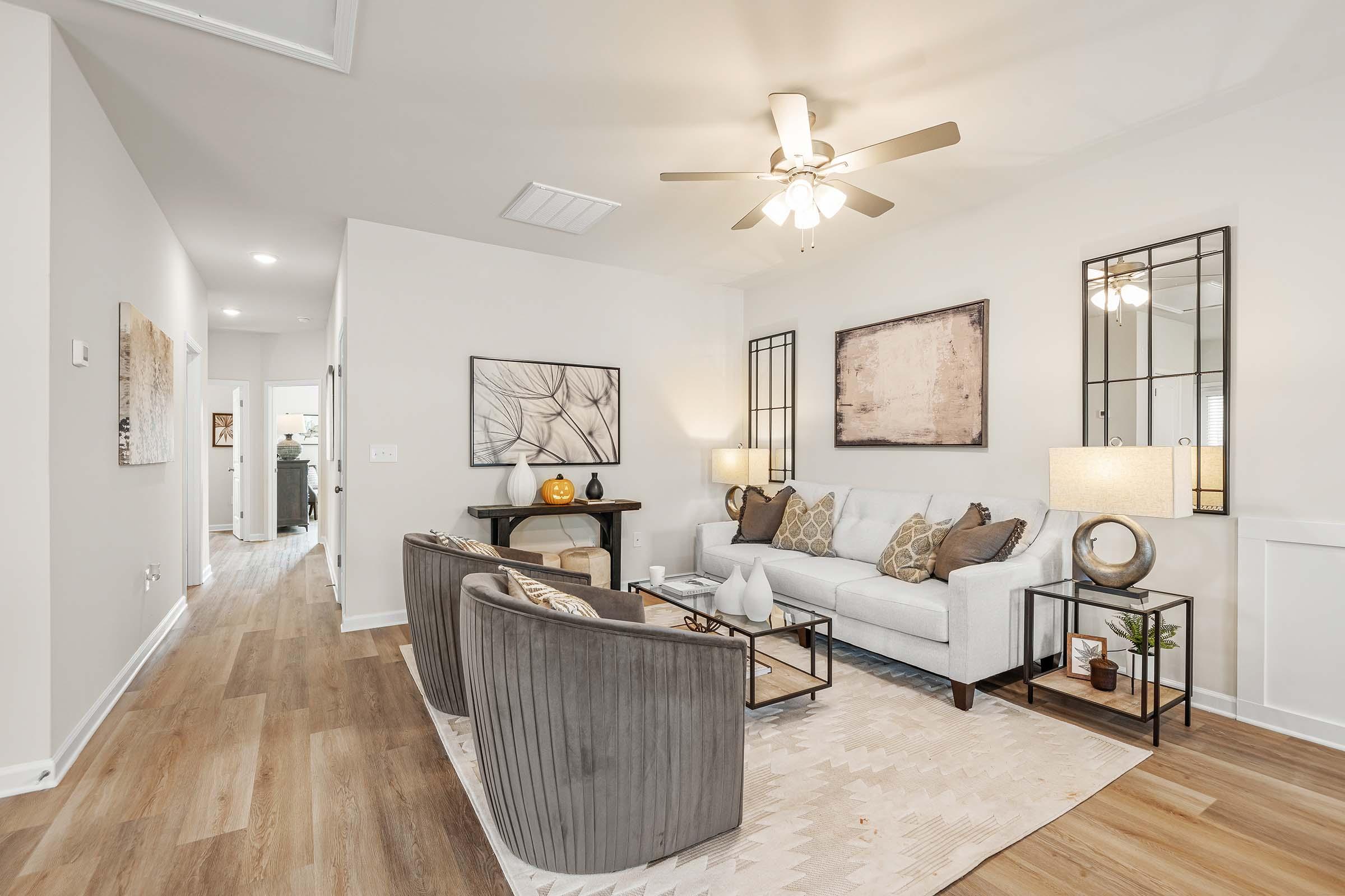 A cozy living room featuring a light gray sofa with decorative pillows, two stylish accent chairs, a coffee table, and a patterned area rug. The space has a ceiling fan, wall art, and mirrors, with warm lighting creating an inviting atmosphere. A hallway is visible in the background.