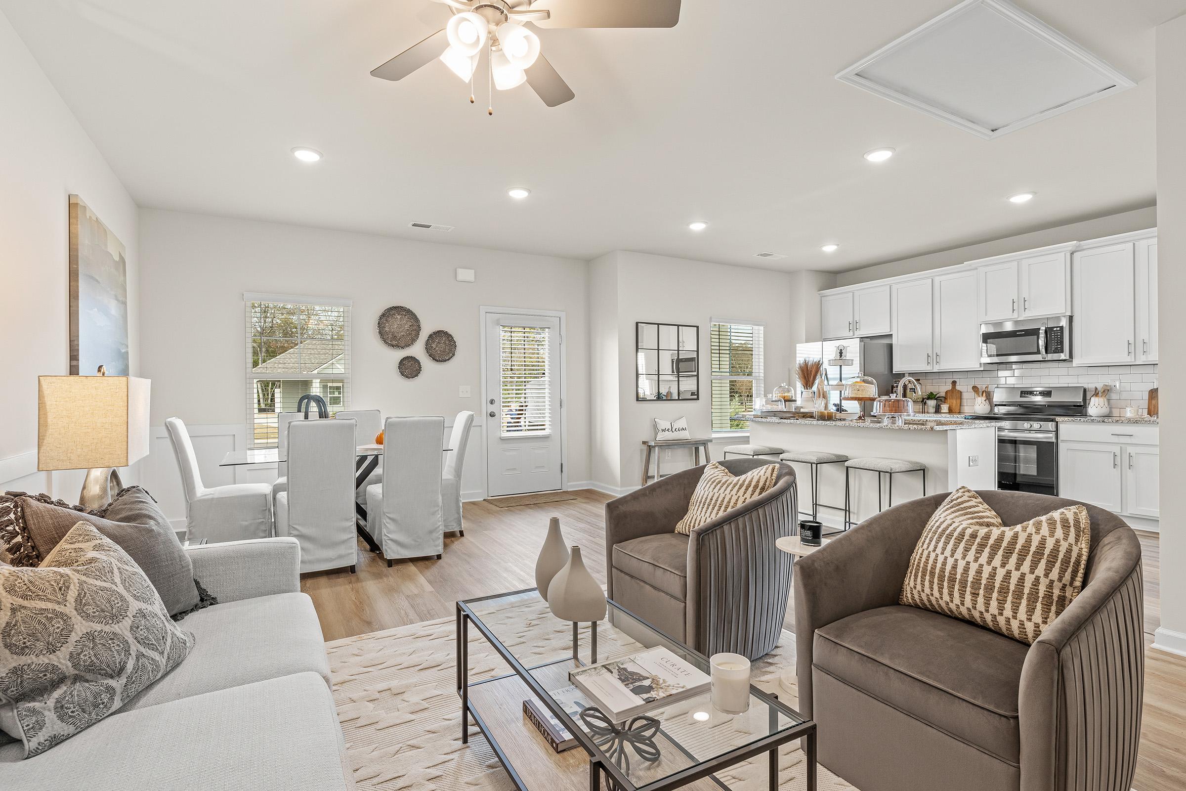 Modern living room and dining area featuring a cozy sofa, two armchairs, a coffee table, and a dining set. The space is bright with natural light, showcasing neutral tones and contemporary decor. A ceiling fan adds comfort, and large windows provide views of the outside.