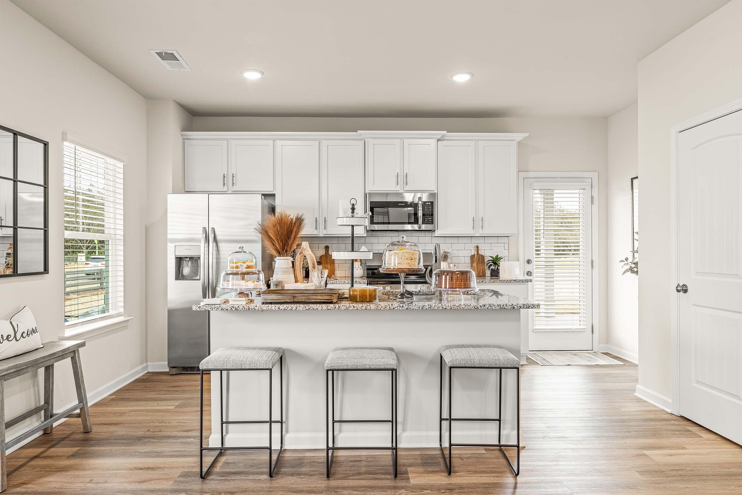 Modern kitchen featuring white cabinetry, stainless steel appliances, and a spacious island with three bar stools. The countertop displays decorative items and a small plant. Large windows allow natural light, and a door leads to an outdoor space. The flooring is wood-like, adding warmth to the design.