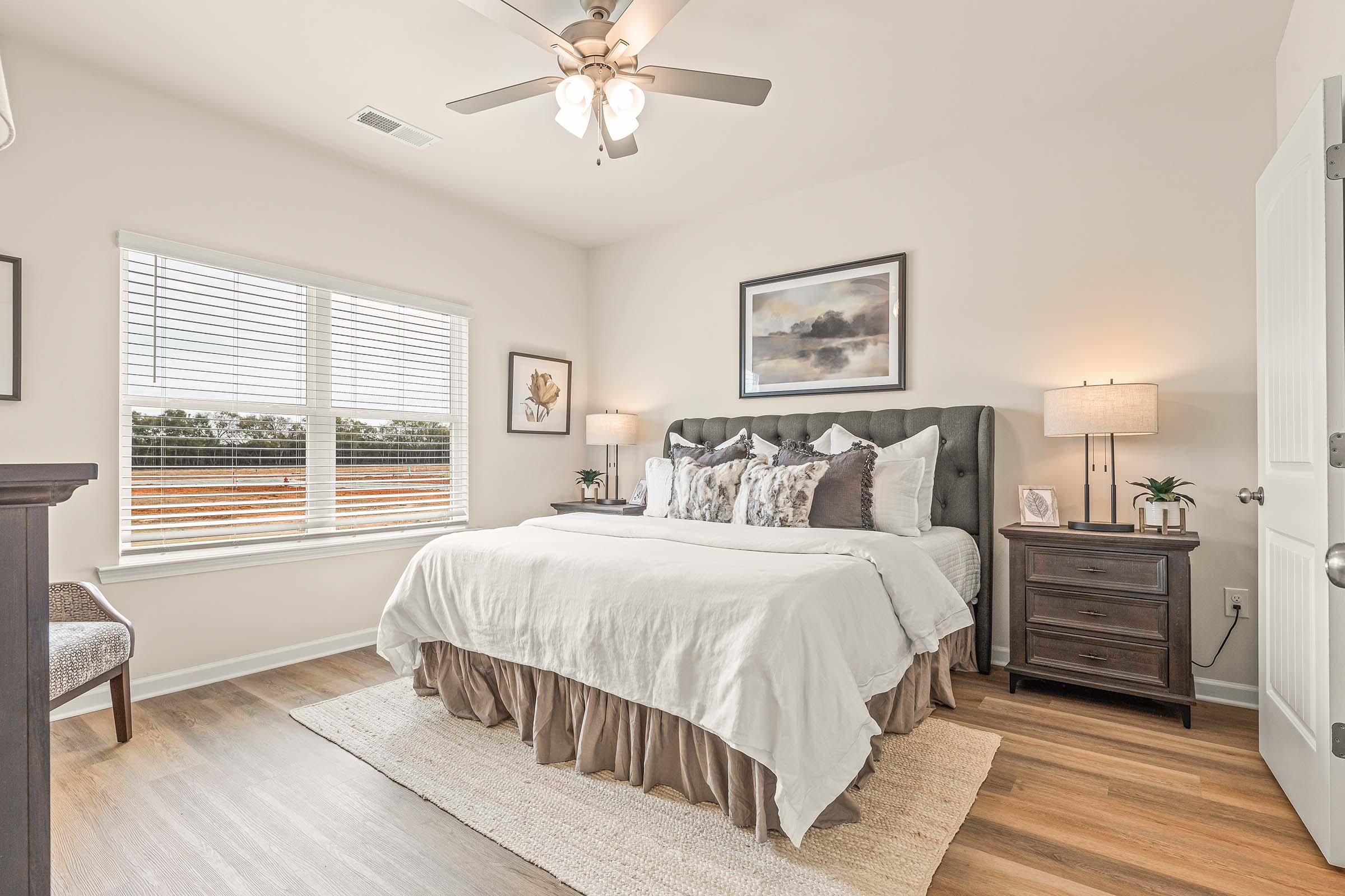 A modern bedroom featuring a queen-sized bed with decorative pillows, a wooden nightstand with a lamp, and a cozy armchair. Natural light streams in through a window with horizontal blinds, and a textured rug adds warmth to the space. Wall art and a ceiling fan complete the inviting atmosphere.