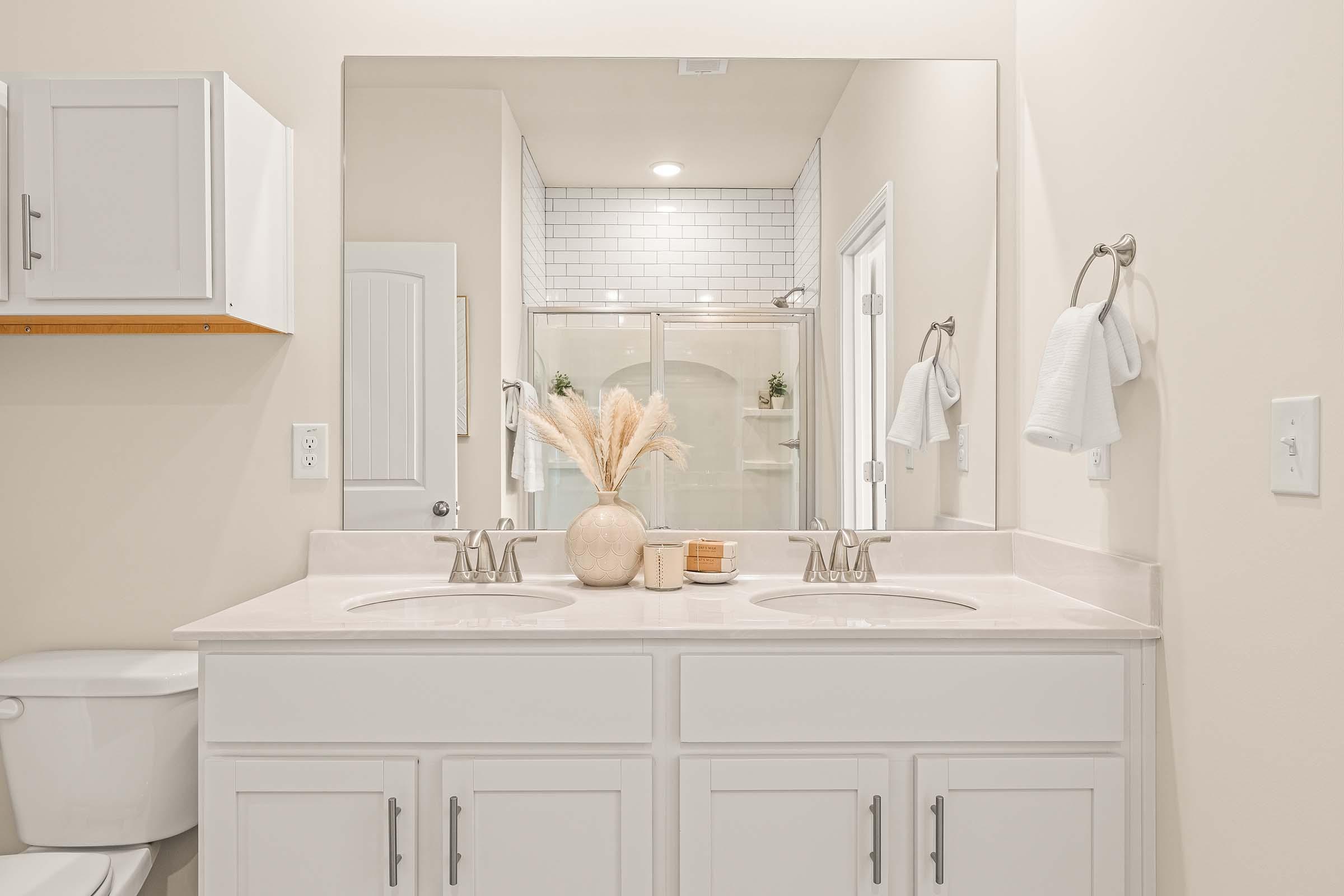 A modern bathroom featuring a double sink vanity with white cabinets, a large mirror, and decorative elements like dried plants and candles. In the background, there's a glass shower and white towels hanging on a towel rack. The overall color scheme is neutral and clean.