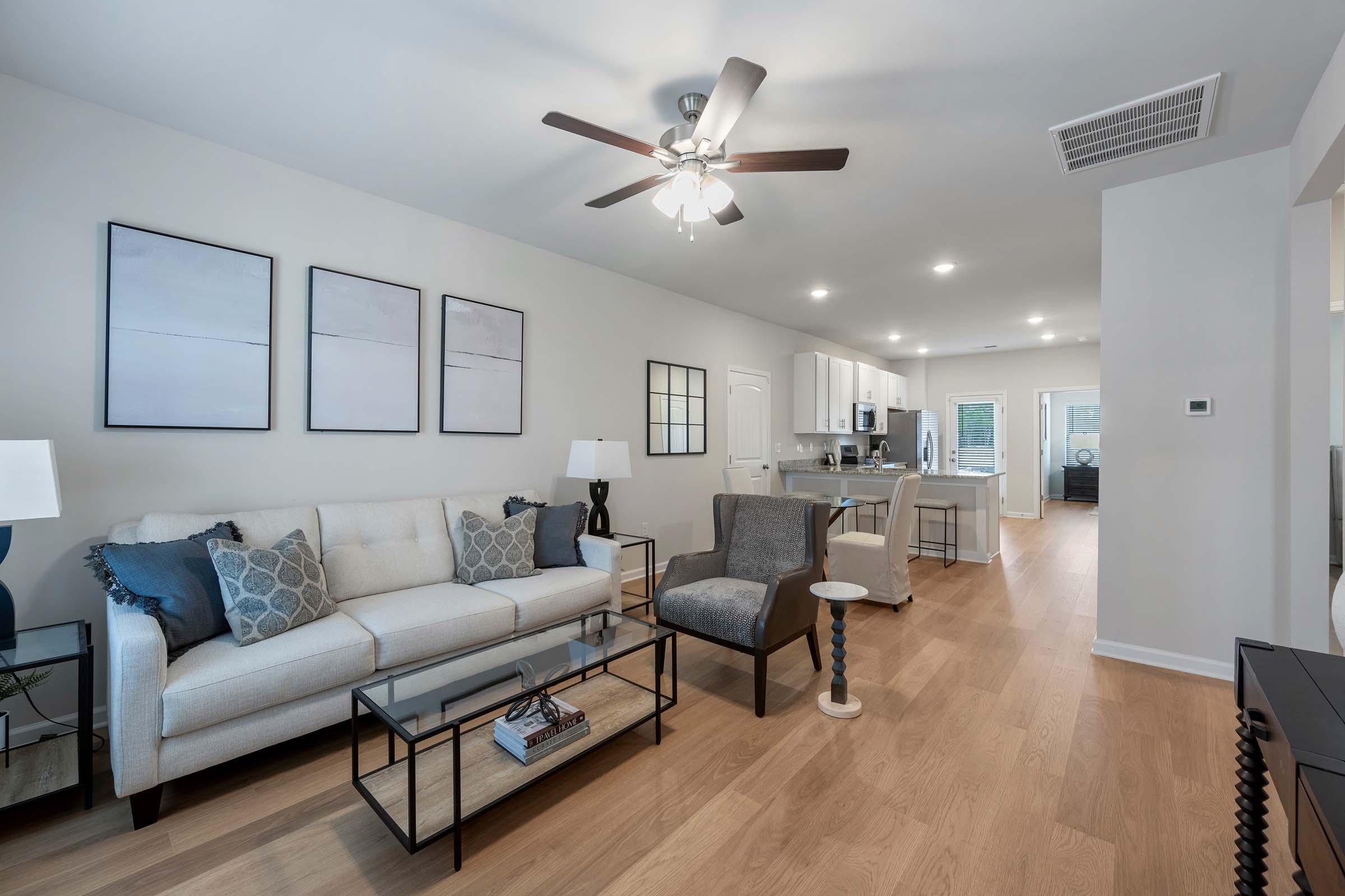 A modern living room featuring a light-colored sofa with decorative pillows, a glass coffee table, and a stylish accent chair. The walls are adorned with three framed artworks, and there's a ceiling fan. The space has an open layout with a view into the dining and kitchen area, illuminated by natural light.