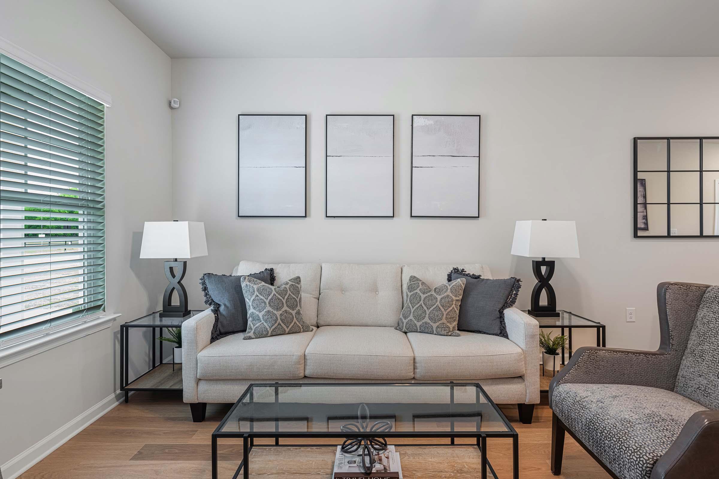 A stylish living room featuring a light-colored sofa with decorative pillows, two modern lamps on side tables, and a glass coffee table. The wall is adorned with three minimalist framed artworks. A cozy armchair is positioned in the corner, and natural light streams in through a window with green blinds.