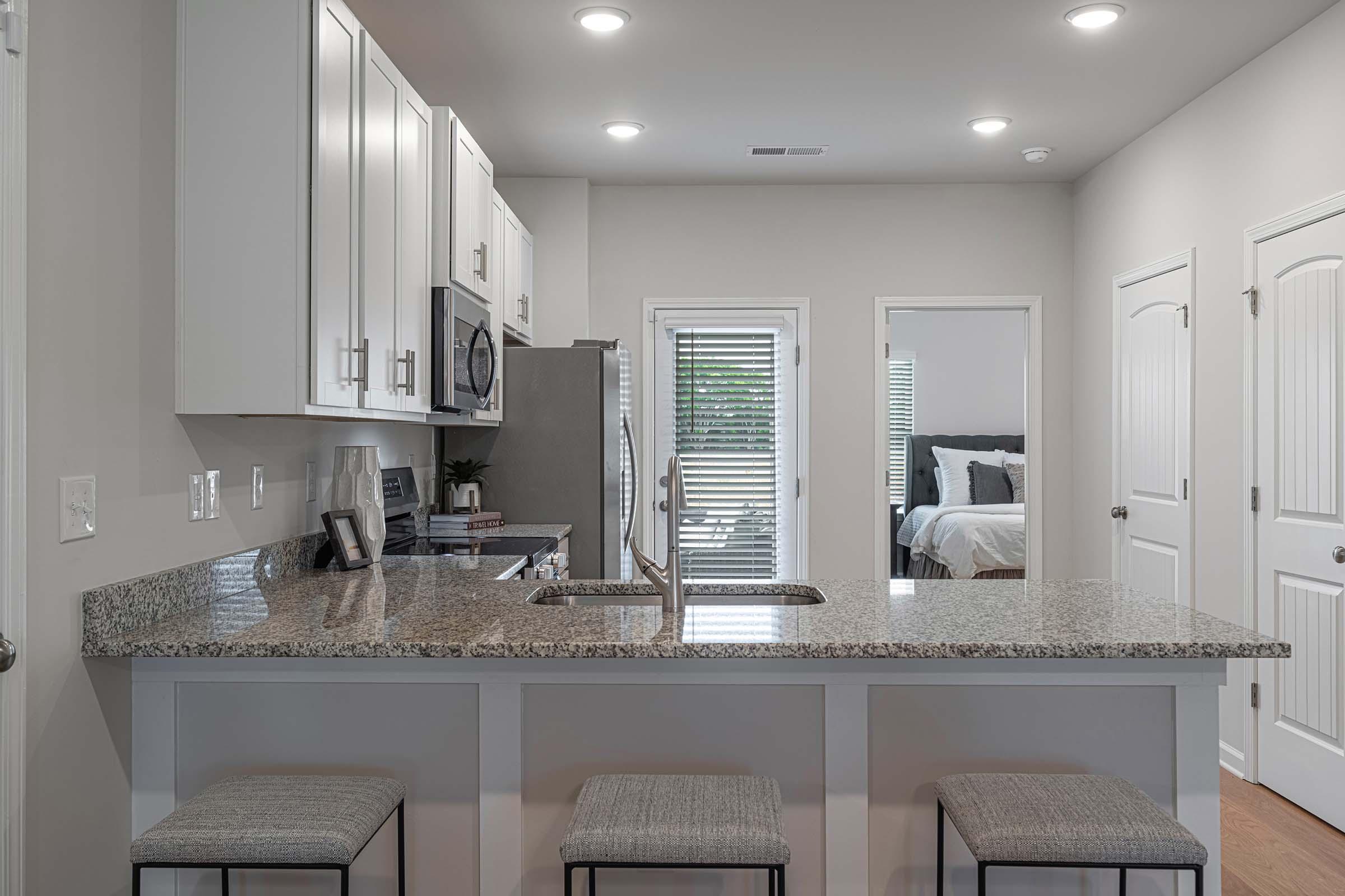 Modern kitchen featuring granite countertops, stainless steel appliances, and white cabinets. A sleek bar area with stools is visible, and there's a door leading to another room in the background. Natural light enters through a window with blinds, creating a bright and inviting atmosphere.