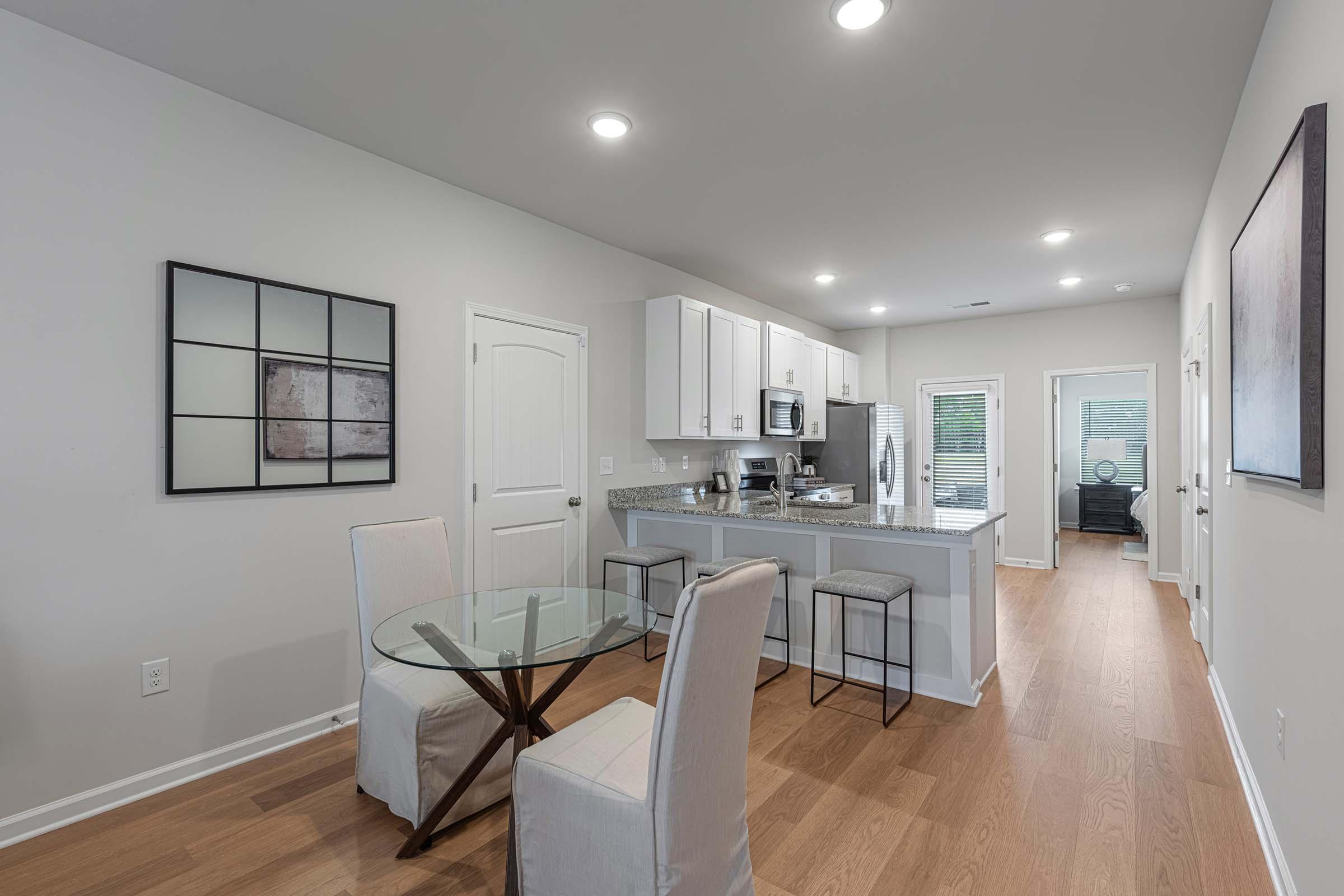 A modern kitchen and dining area featuring a glass-top dining table with four chairs, stainless steel appliances, and white cabinetry. The space has hardwood flooring, recessed lighting, and large windows with blinds, creating a bright and airy atmosphere.