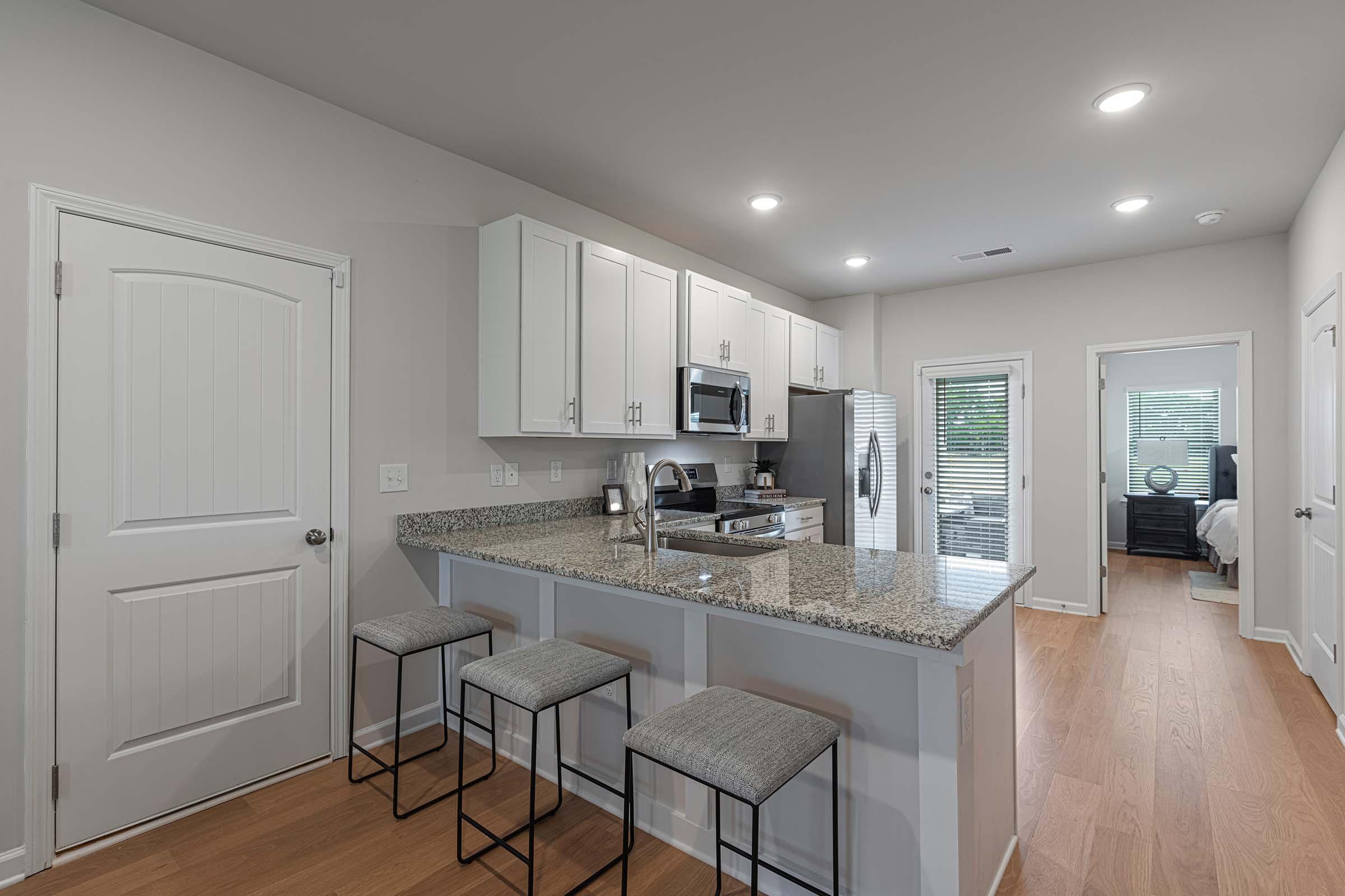 Modern kitchen featuring granite countertops, stainless steel appliances, and white cabinetry. There are three bar stools at the counter, and a doorway leading to a bedroom area is visible in the background. The space is well-lit with recessed lighting and has a clean, contemporary design.