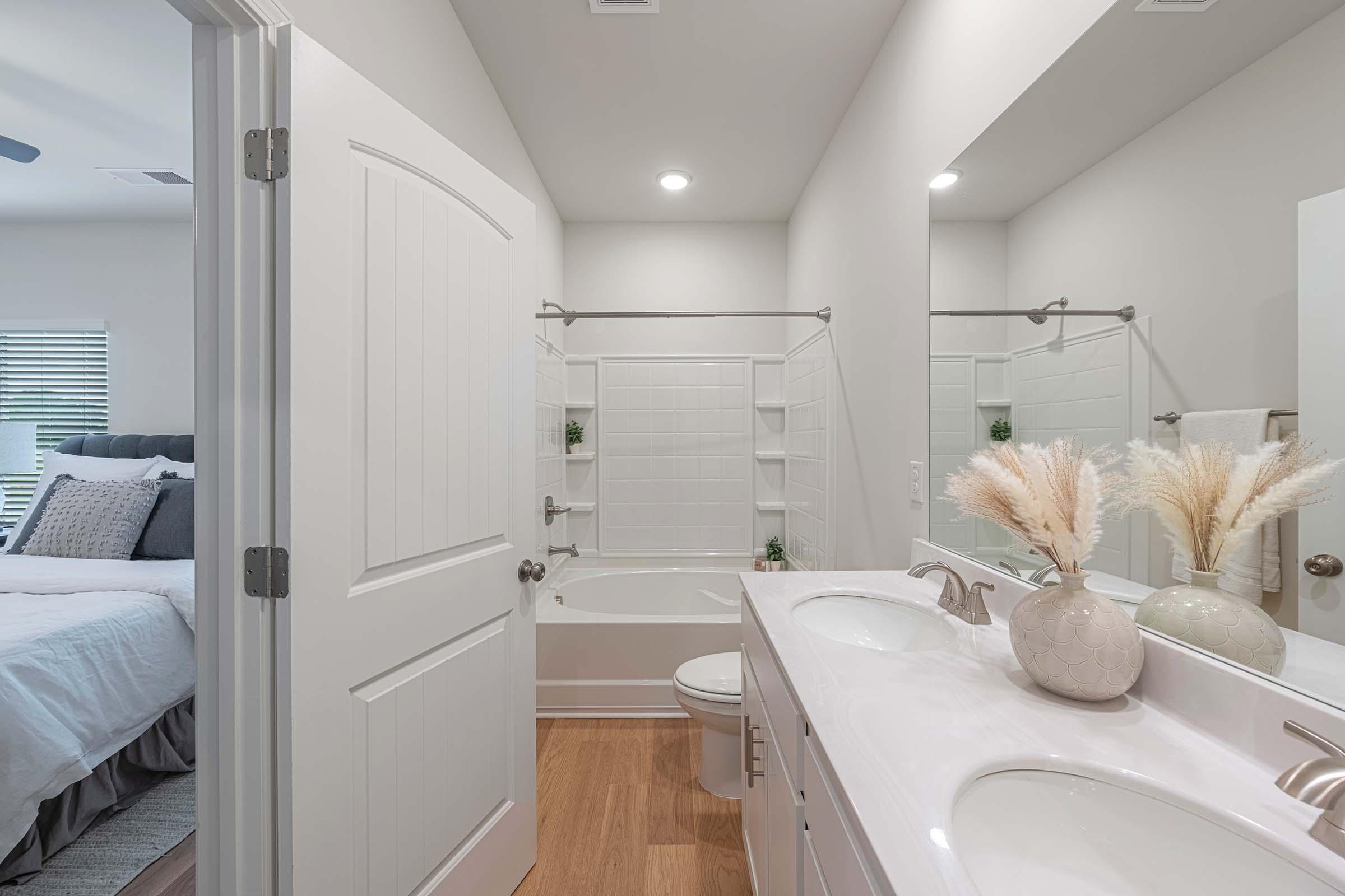 A modern bathroom featuring a white bathtub and shower combination, a double vanity with light-colored countertops, and decorative vases with dried flowers. The space is well-lit with overhead lights and has a neutral color scheme, complemented by a glimpse of a bedroom through the doorway.