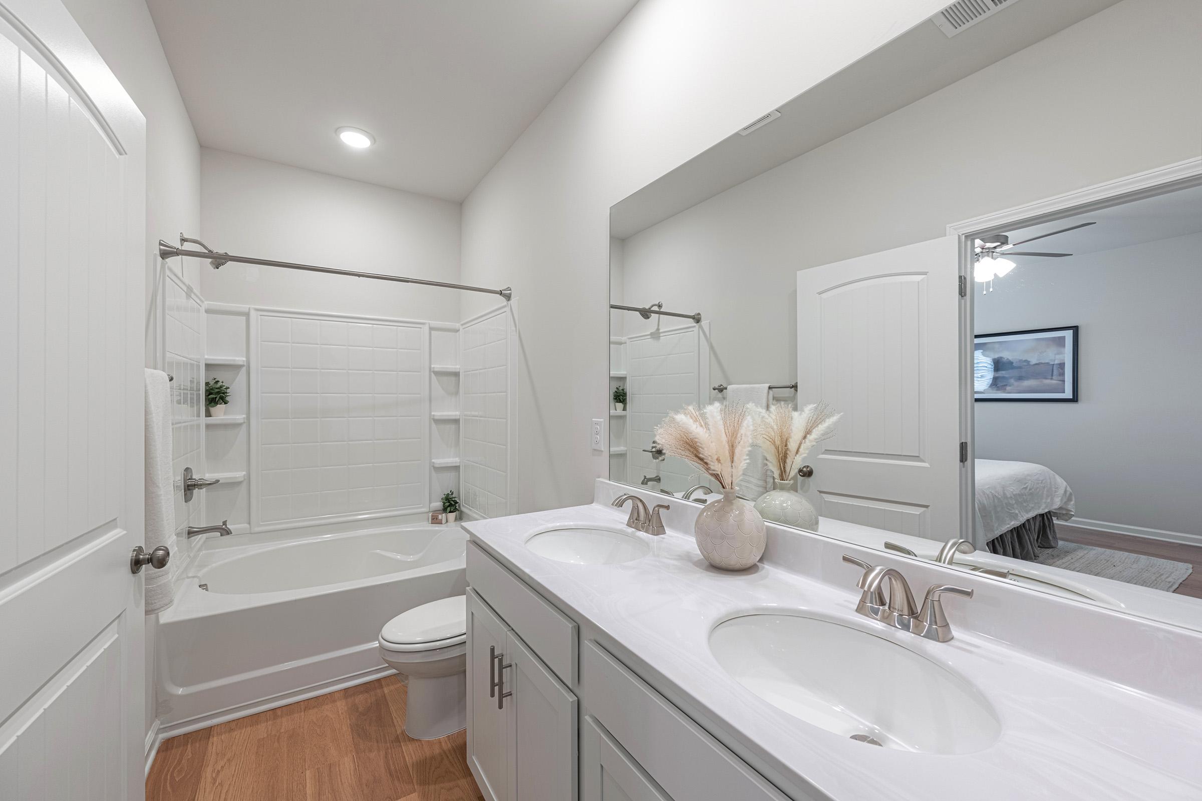A modern bathroom featuring a clean, white design with a bathtub and shower combination. Two sinks are set in a light-colored countertop, and decorative plants add a touch of elegance. Natural light illuminates the space, creating a bright and inviting atmosphere. A mirror reflects the overall aesthetic.