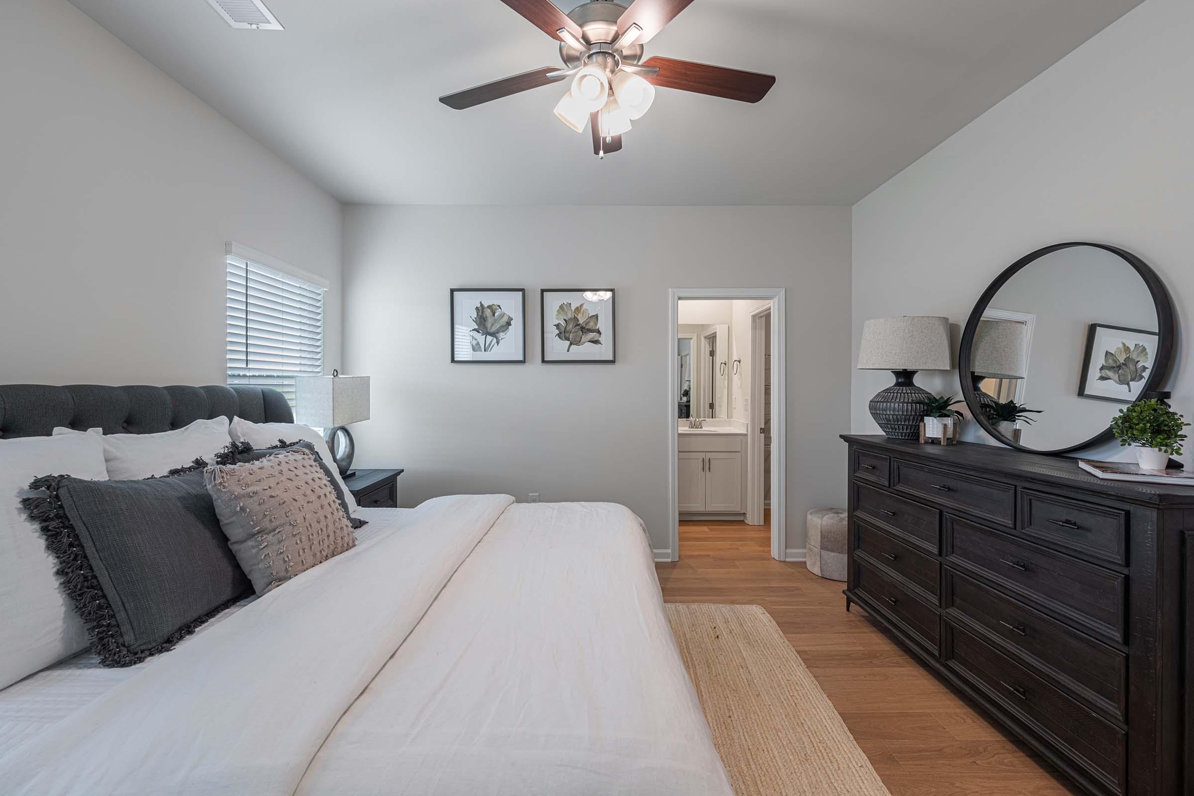 A stylish bedroom featuring a large bed with white bedding and decorative pillows. There's a dresser, a round mirror, and framed botanical prints on the wall. The room has a ceiling fan and warm wooden flooring, creating a cozy atmosphere. A bathroom entrance is visible in the background.