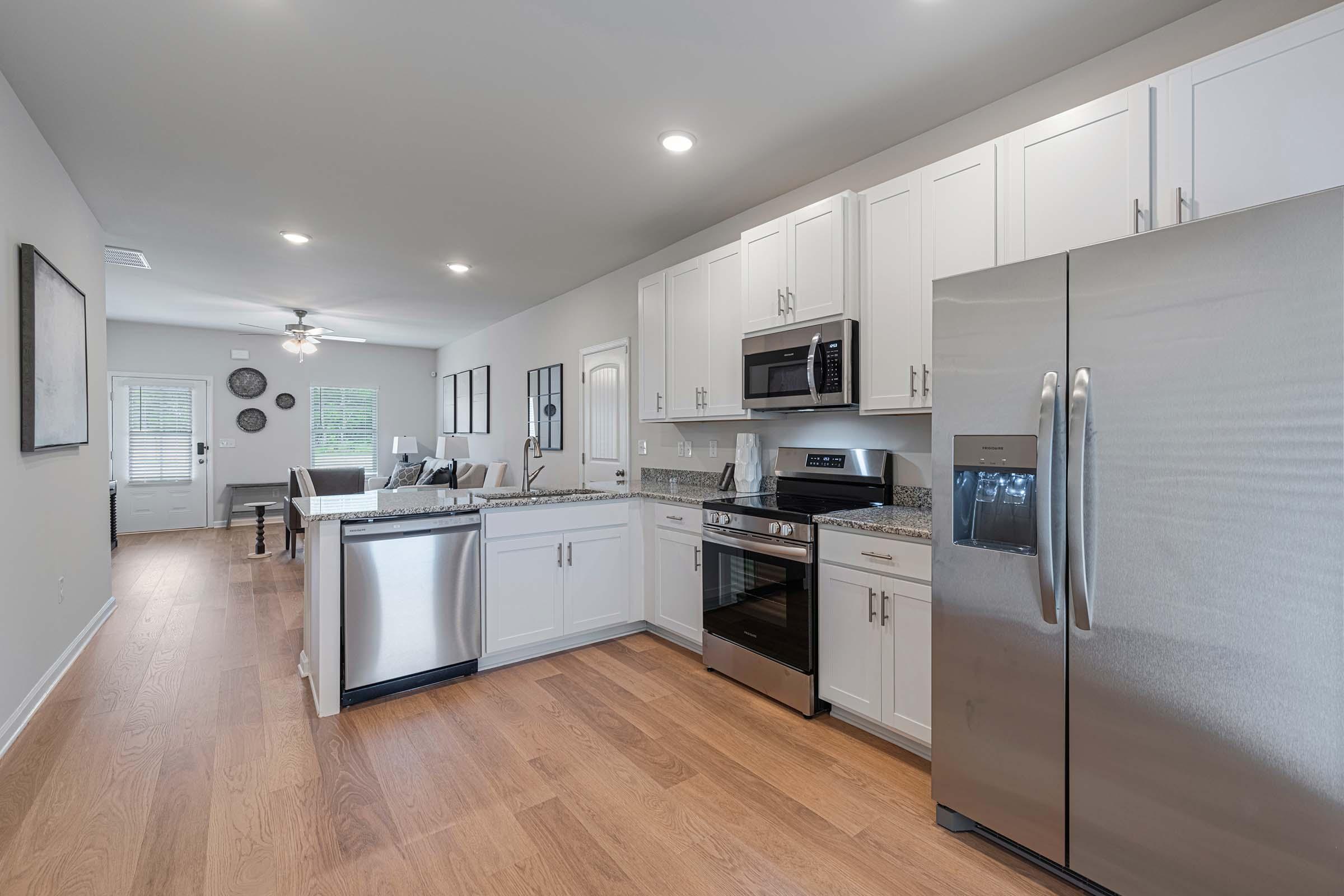 Modern kitchen featuring white cabinets, stainless steel appliances including a refrigerator and oven, granite countertops, and hardwood floors. The layout includes a dining area with a ceiling fan and natural light from windows, creating a bright and inviting space.
