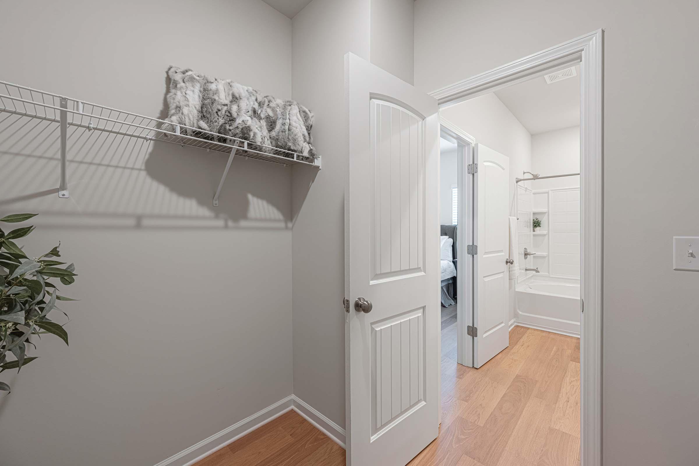 A view of a modern, light-colored bathroom entrance with an open door. There is a towel rack with folded towels on the left wall and a plant nearby. The bathroom features a white bathtub and shower combination, with gray walls and wooden flooring visible in the foreground.