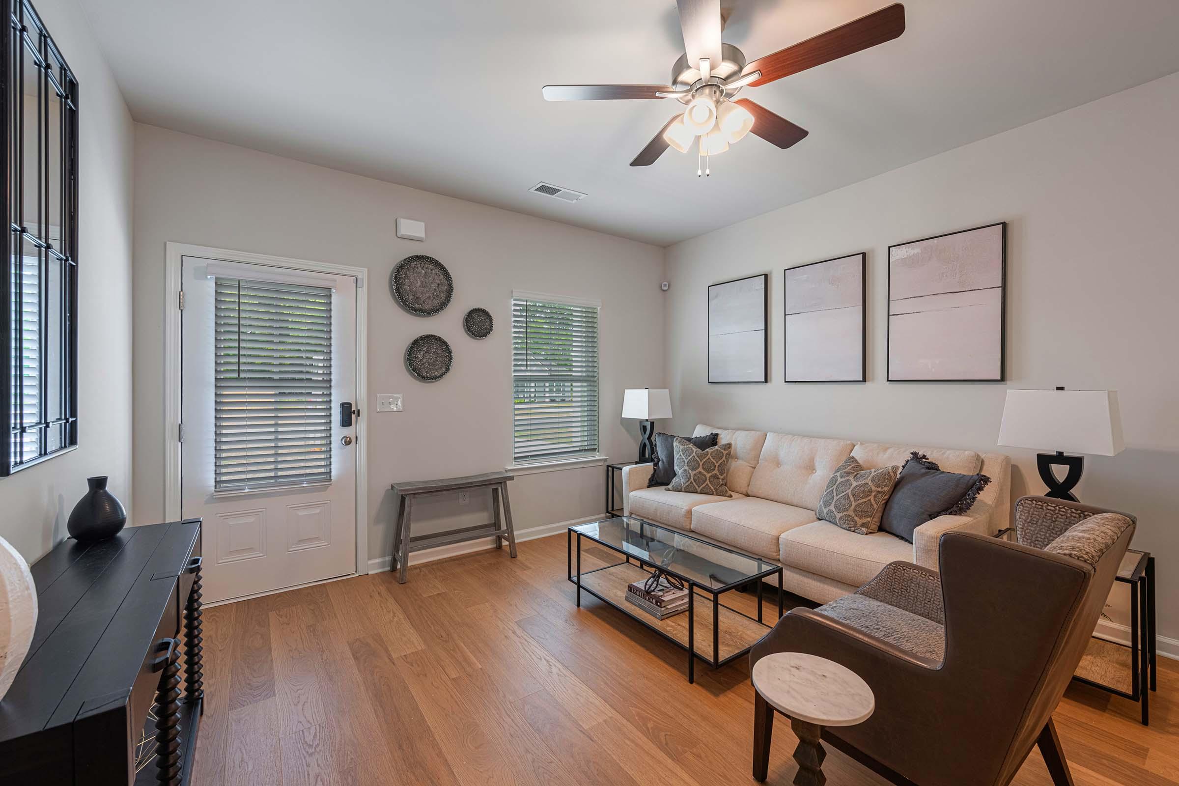 A cozy living room featuring a beige sofa with decorative pillows, a glass coffee table, and a dark wood side table. There are two lamps on either side of the sofa and three framed artworks on the wall. A door leads outside, and there are round wall decorations above the sofa. The flooring is hardwood.